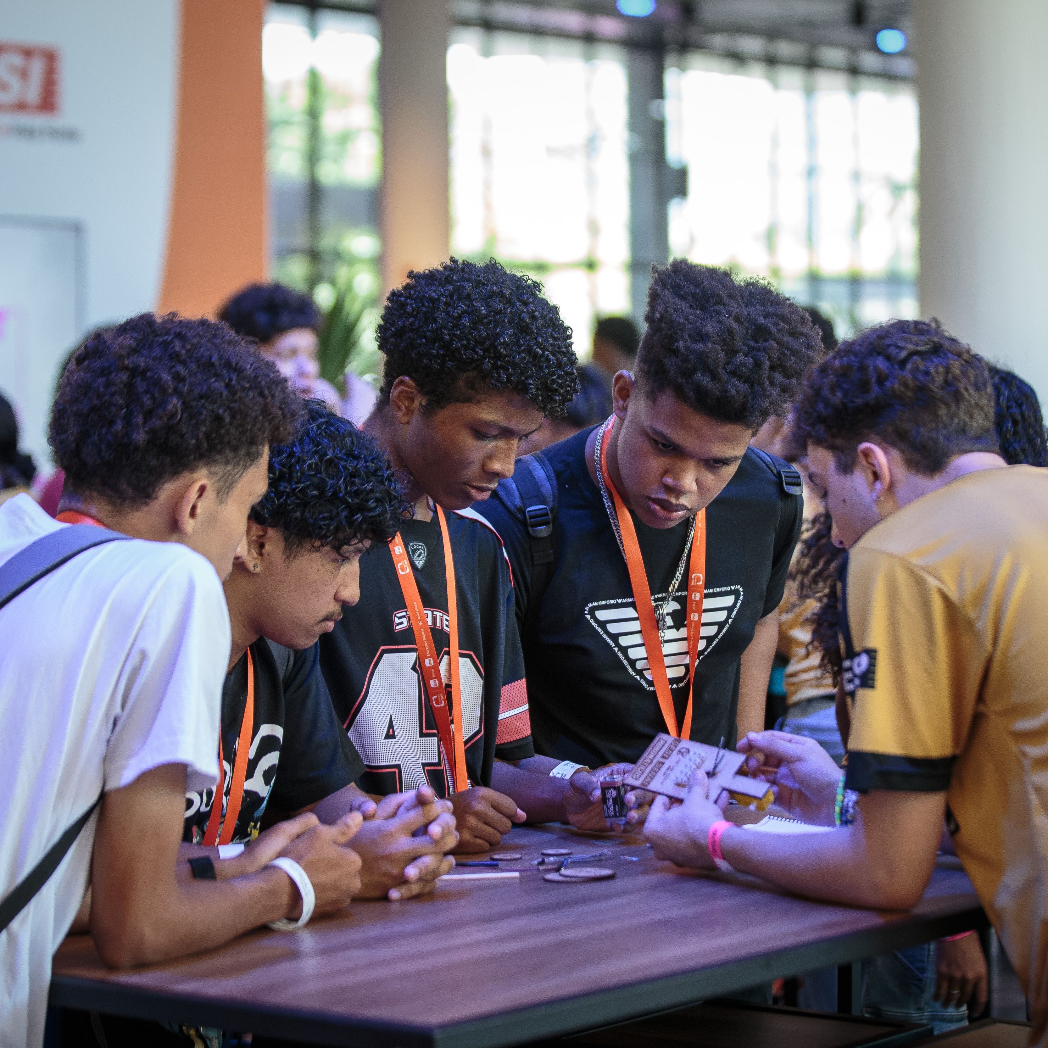 Fotografia a cores na horizontal que mostra um grupo de jovens rapazes reunidos &agrave; volta de uma mesa de madeira num pavilh&atilde;o amplo e bem iluminado. Eles observam com muita aten&ccedil;&atilde;o um pequeno objeto que est&aacute; a ser manuseado pelo jovem posicionado &agrave; direita.   O rapaz &agrave; direita est&aacute; de costas para a c&acirc;mara, veste uma t-shirt amarela e preta com v&aacute;rios log&oacute;tipos nas costas e a inscri&ccedil;&atilde;o 