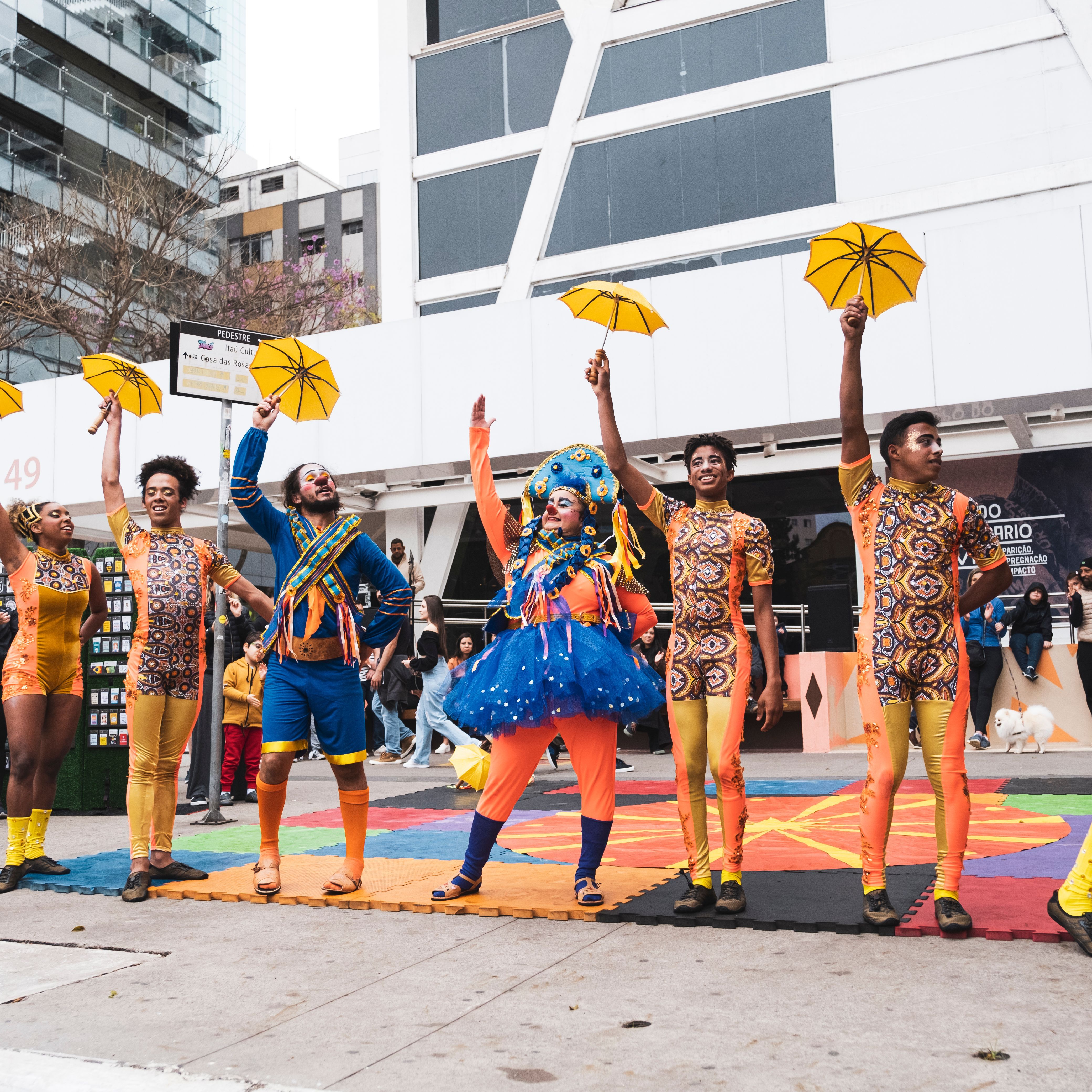 A fotografia mostra uma apresenta&ccedil;&atilde;o de rua ao ar livre, em frente ao pr&eacute;dio do Ita&uacute; Cultural. No centro, um artista equilibra-se em um monociclo alto, interagindo com o p&uacute;blico. Ele veste um casaco vinho e bermuda. Uma grande roda de pessoas, formada por adultos e crian&ccedil;as, assiste &agrave; performance com aten&ccedil;&atilde;o. Algumas crian&ccedil;as est&atilde;o sentadas no ch&atilde;o na primeira fileira, enquanto v&aacute;rias pessoas em p&eacute; tiram fotos com seus celulares.   Ao fundo, a fachada branca do edif&iacute;cio exibe um cartaz marrom da exposi&ccedil;&atilde;o 