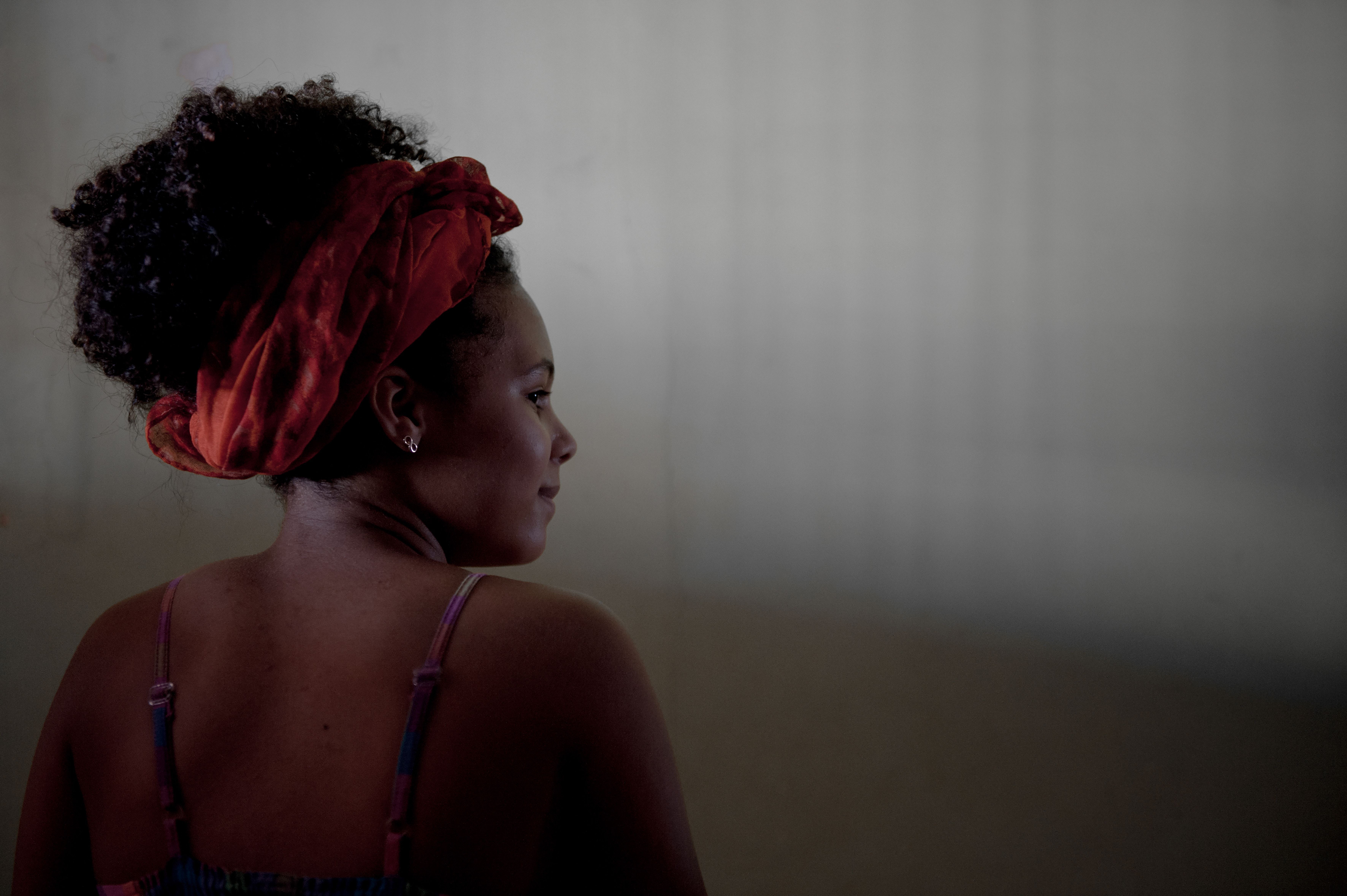 Uma mulher negra aparece de costas, com o rosto de perfil, olhando para a direita. Os seus cabelos est&atilde;o presos por um len&ccedil;o vermelho, que se destaca na imagem. A ilumina&ccedil;&atilde;o suave e o fundo desfocado criam uma atmosfera intimista e contemplativa.