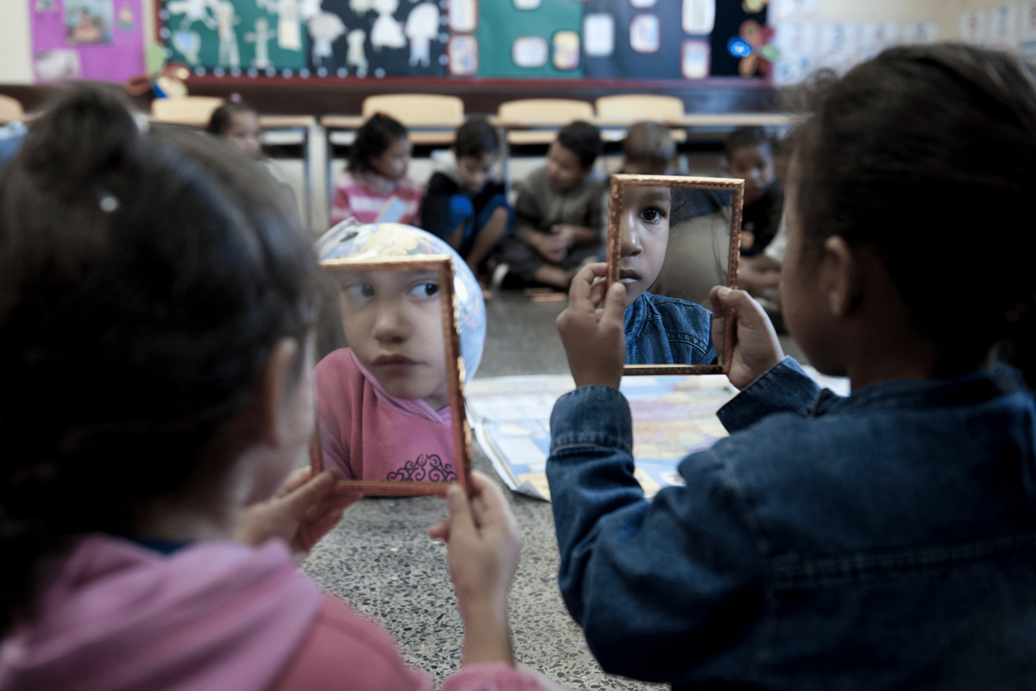 Duas crian&ccedil;as est&atilde;o sentadas no ch&atilde;o de uma sala de aula, segurando pequenos espelhos retangulares com molduras douradas. Nos espelhos, aparecem os reflexos de seus rostos, enquanto um globo terrestre e pap&eacute;is coloridos est&atilde;o sobre o ch&atilde;o. Ao fundo, outras crian&ccedil;as est&atilde;o sentadas em grupo, diante de mesas, cadeiras e murais coloridos nas paredes.