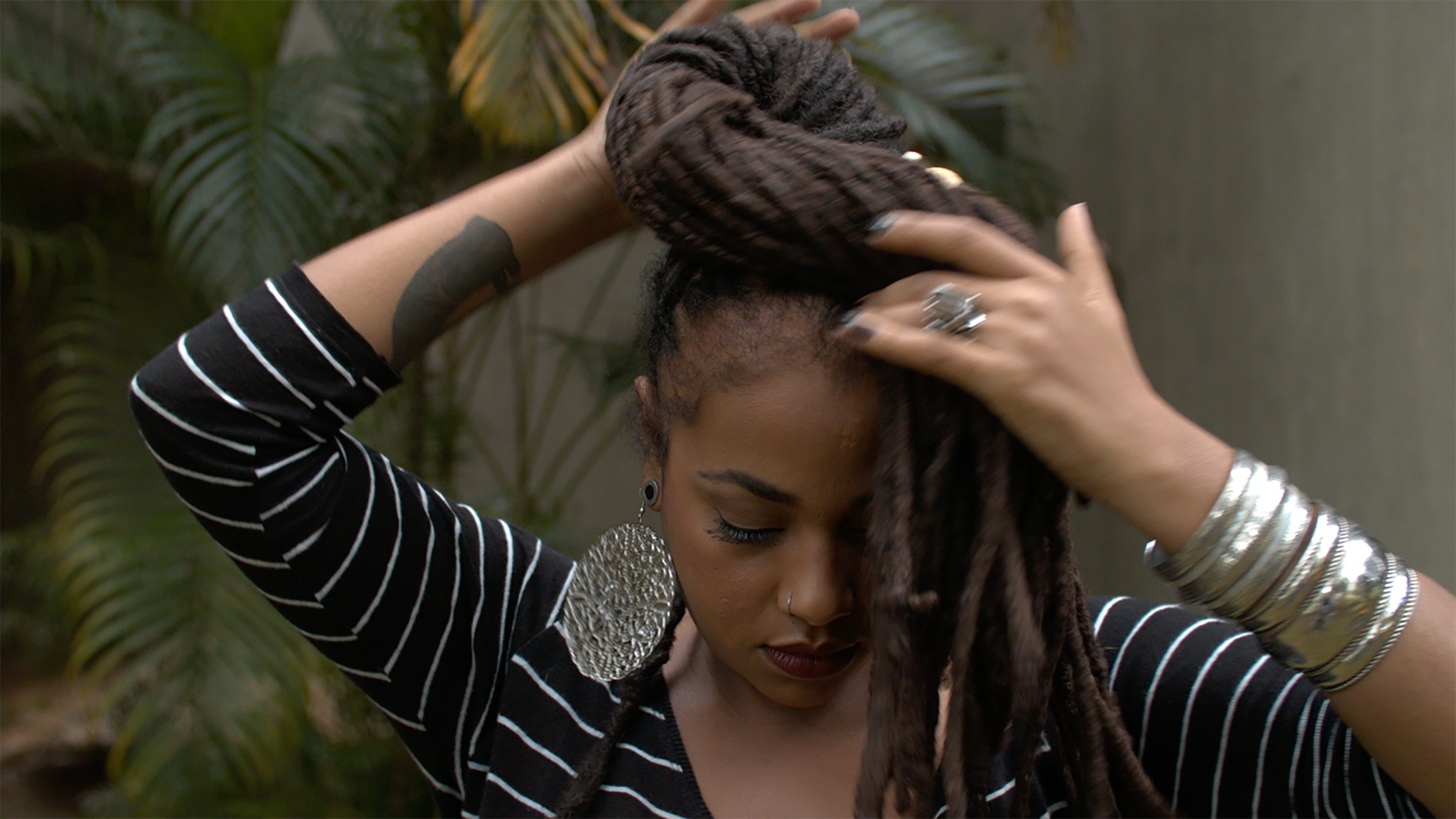 Uma mulher negra, com a cabe&ccedil;a inclinada para baixo, segura seus cabelos longos e tran&ccedil;ados com as duas m&atilde;os. Ela veste uma blusa preta com listras brancas, brincos grandes prateados, an&eacute;is e v&aacute;rias pulseiras met&aacute;licas. Ao fundo, aparecem plantas de folhas verdes e uma parede em tons de cinza.