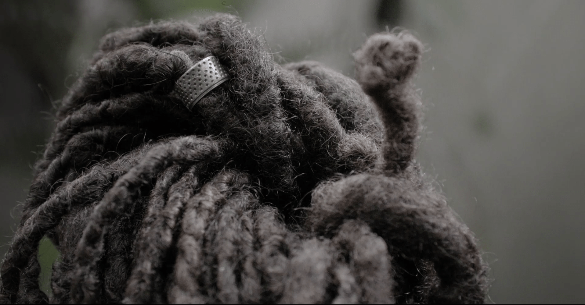 A imagem mostra um close de cabelos escuros, volumosos e organizados em mechas grossas ou dreadlocks. Algumas mechas est&atilde;o presas no alto da cabe&ccedil;a por um acess&oacute;rio met&aacute;lico prateado com pequenos furos.