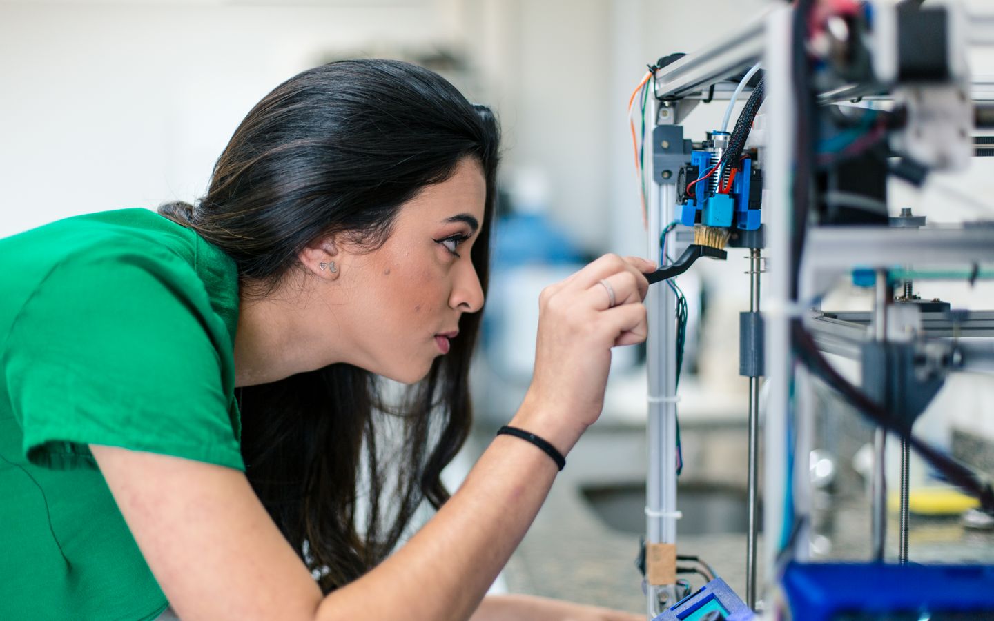 Na imagem, há uma menina branca com longos cabelos lisos e pretos. Ela está vestindo uma camisa verde e tem uma expressão séria no rosto. A menina está olhando em direção a alguns equipamentos tecnológicos enquanto realiza a manutenção deles.