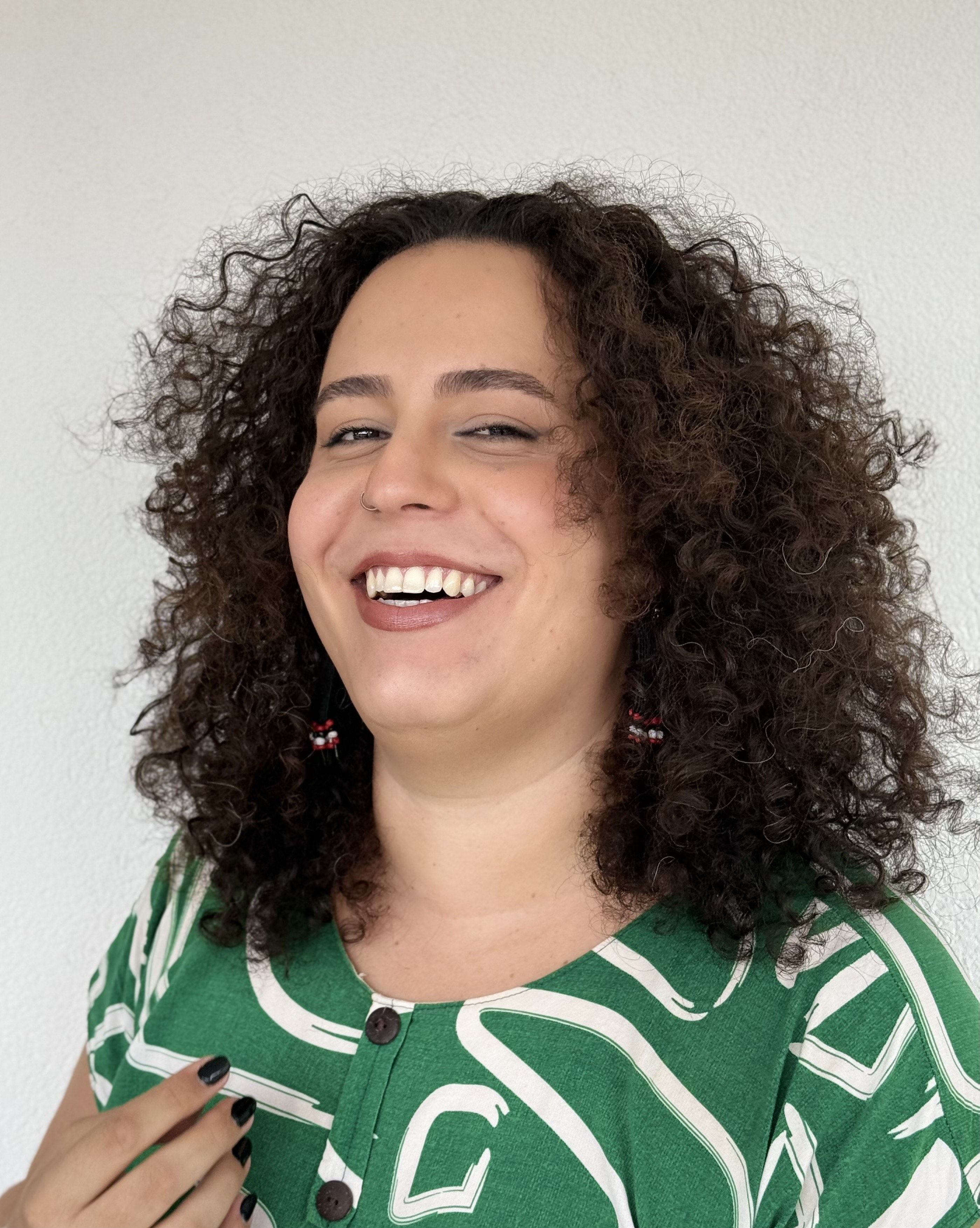 Retrato da professora e pesquisadora Era Grey. Ela é uma mulher branca, de cabelos encaracolados e sorri para a câmera. Ela veste uma blusa verde com grafismos em branco.