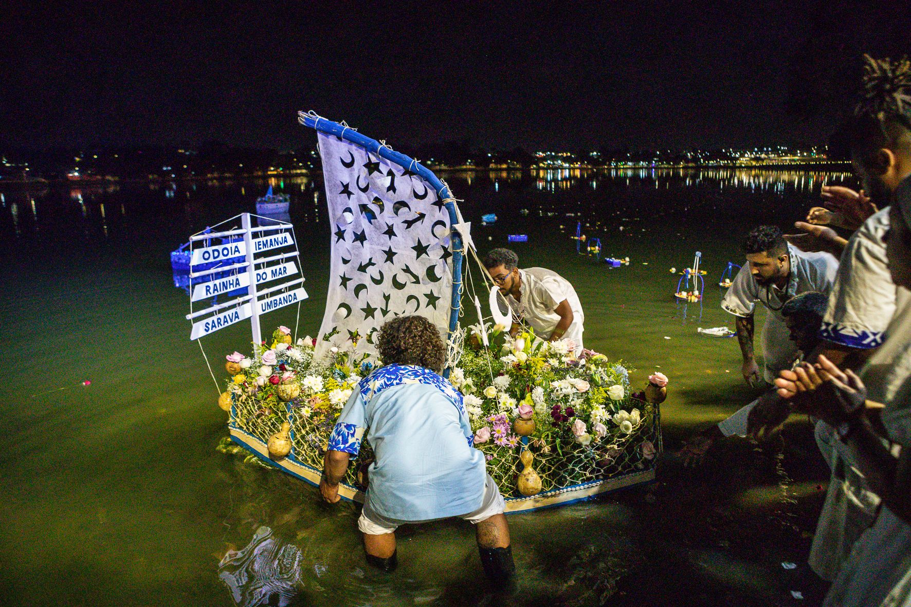 Fotografia colorida de integrantes da Casa de Caridade Pai Jacob do Oriente colocando um barco para Iemanja na Lagoa da Pampulha. O barco é repleto de flores e tem uma vela de pano branca presa em um mastro azul.