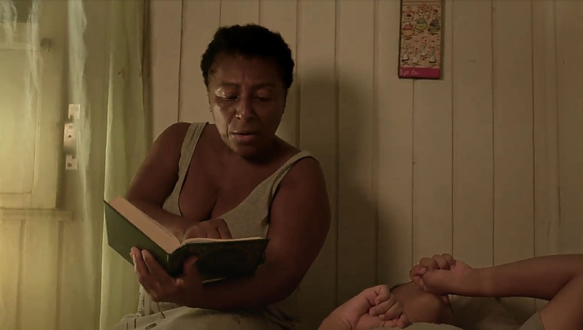 Nininha está lendo um livro para o filho. Ela é uma senhora negra e está vestindo uma camisola cinza. O menino está deitado na cama, ao lado de Nininha.