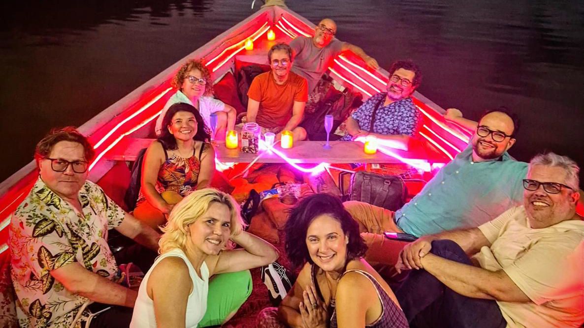 
Sete adultos sorridentes em um barco iluminado com luzes vermelhas e amarelas. Três homens sentam na popa, quatro mulheres e dois homens sentam no meio, e um homem senta na proa.