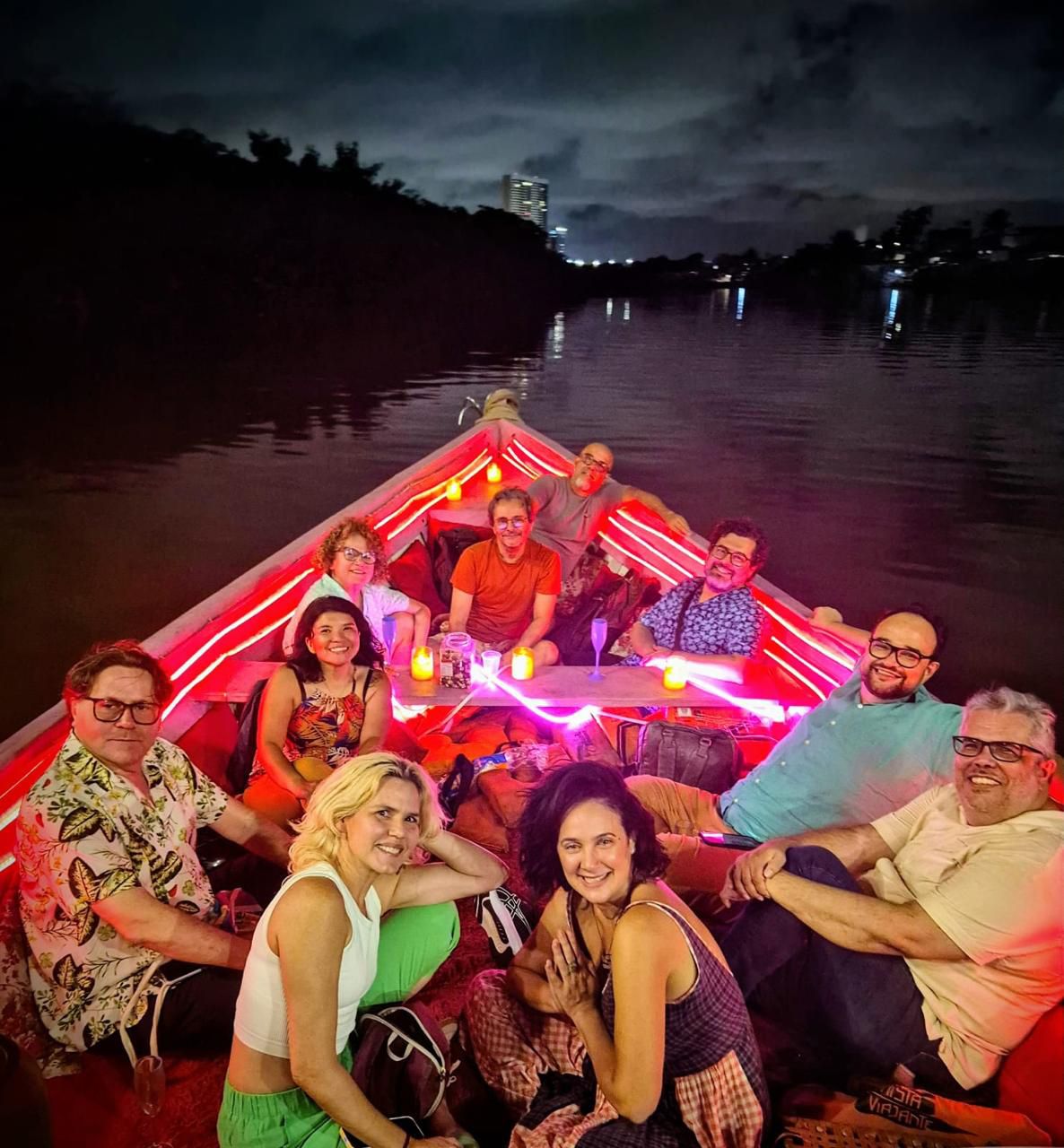 
Sete adultos sorridentes em um barco iluminado com luzes vermelhas e amarelas. Três homens sentam na popa, quatro mulheres e dois homens sentam no meio, e um homem senta na proa.