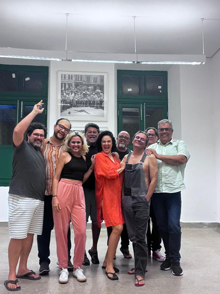 Nove adultos sorridentes em um ambiente interno. Da esquerda para a direita: um homem de barba e camiseta listrada acena; um homem de óculos e camisa listrada; uma mulher loira de top preto e calça rosa; um homem de barba e camiseta preta; uma mulher de vestido vermelho sorri amplamente; um homem careca de óculos e camiseta preta; um homem de óculos escuros e macacão; um homem de óculos e camiseta preta; um homem de óculos e camisa clara. Ao fundo, janelas verdes e um quadro.