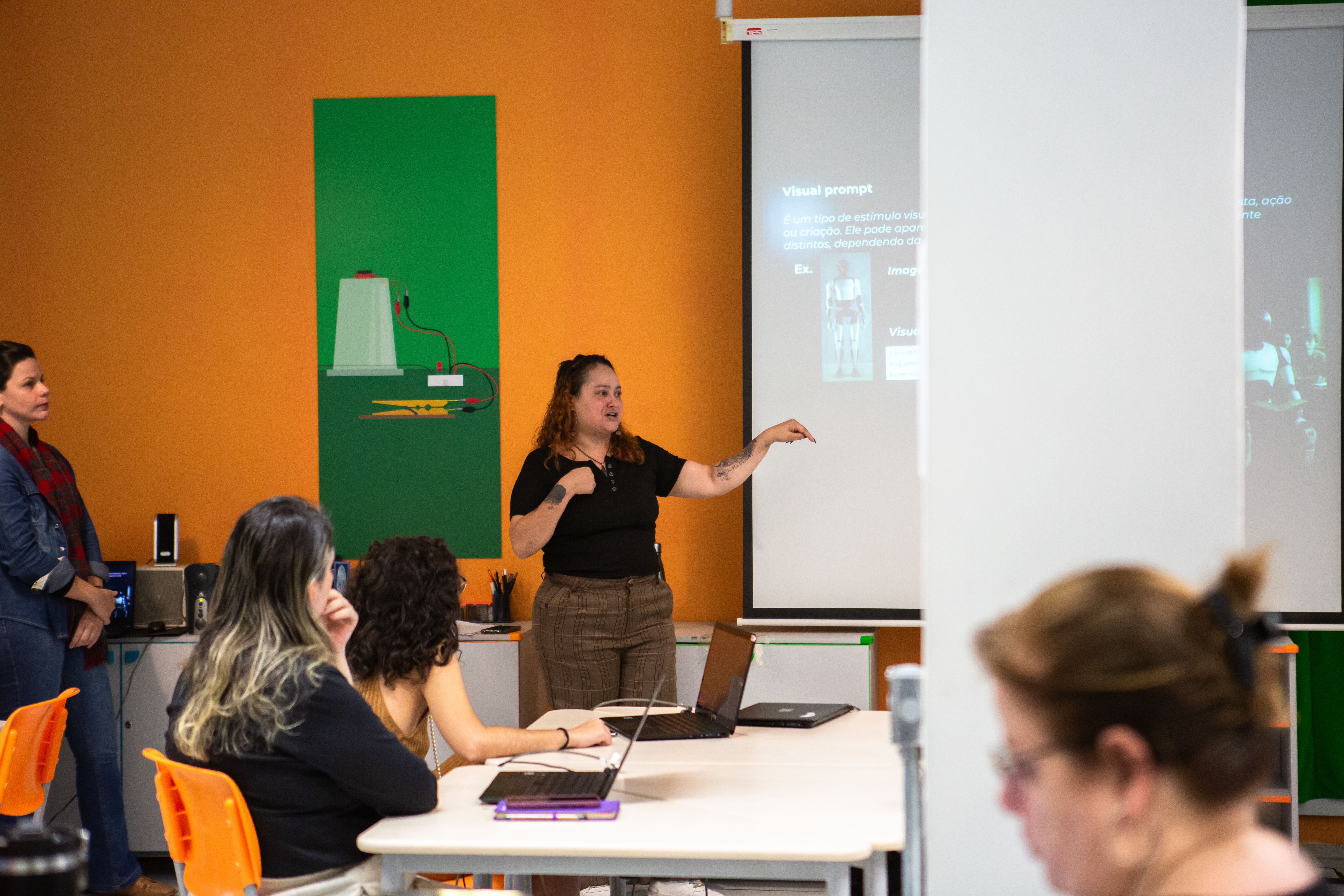 No centro da imagem est&aacute; uma mulher branca, de cabelo ondulado e castanho na altura do ombro,  vestindo camiseta preta e cal&ccedil;a marrom. Ela aponta e dire&ccedil;&atilde;o ao tel&atilde;o onde est&aacute; sendo projetada uma apresenta&ccedil;&atilde;o. A mulher est&aacute; falando e em sua frente est&atilde;o diversas pessoas sentadas falando