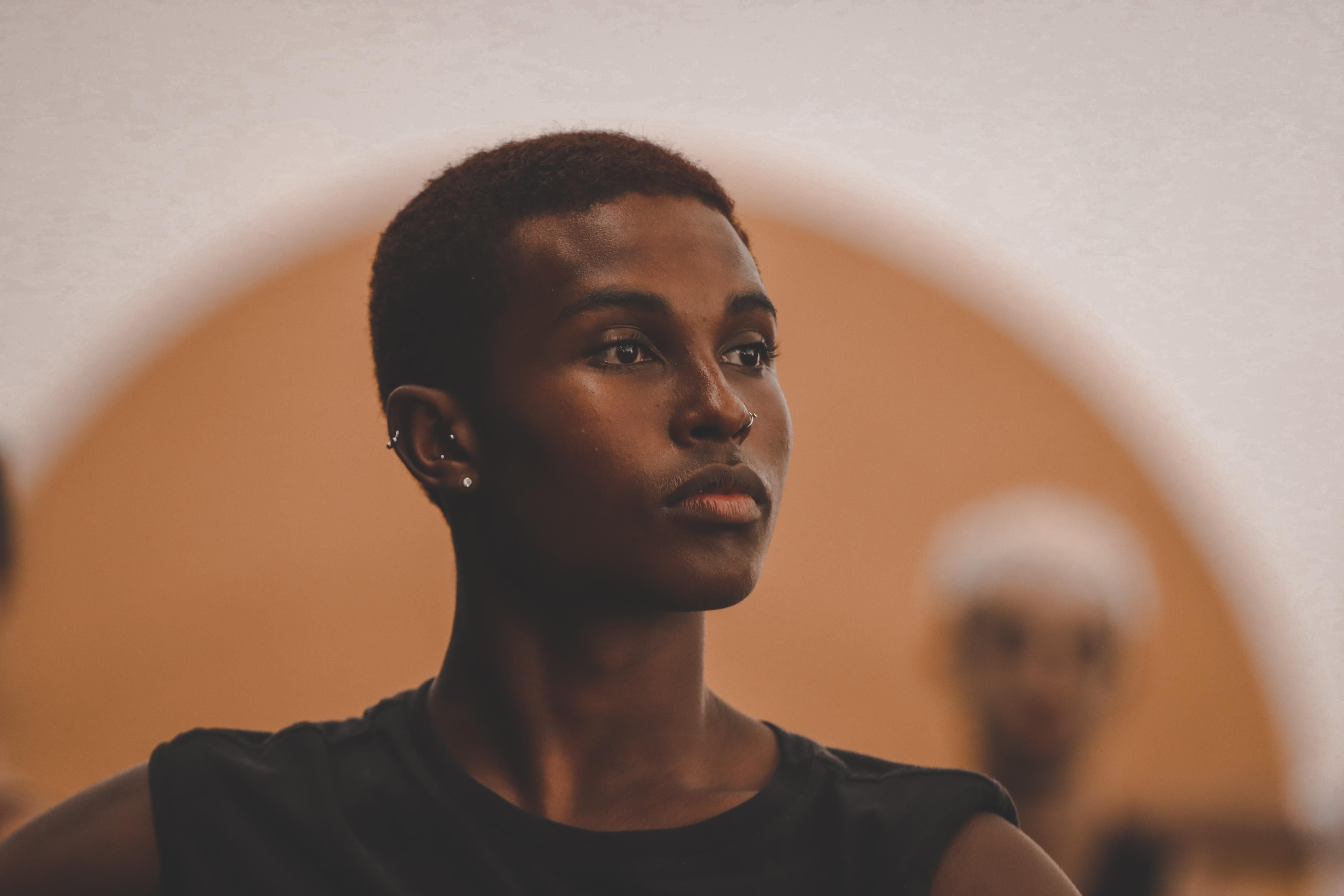 A foto &eacute; um retrato em close-up de uma pessoa jovem de pele escura, com cabelo bem curto e crespo, em um corte que mostra a testa. A pessoa tem um piercing no nariz (argola) e um brinco pequeno na orelha. Os olhos est&atilde;o focados em algo fora do quadro, &agrave; direita, e a express&atilde;o &eacute; s&eacute;ria e contemplativa. A pessoa veste uma camiseta regata preta. O fundo &eacute; suavemente desfocado, com tons de p&ecirc;ssego e um arco mais escuro vis&iacute;vel, sugerindo um ambiente interno, possivelmente um est&uacute;dio de dan&ccedil;a ou teatro, com outra figura desfocada ao fundo &agrave; direita. A ilumina&ccedil;&atilde;o destaca a pele e o perfil do rosto.