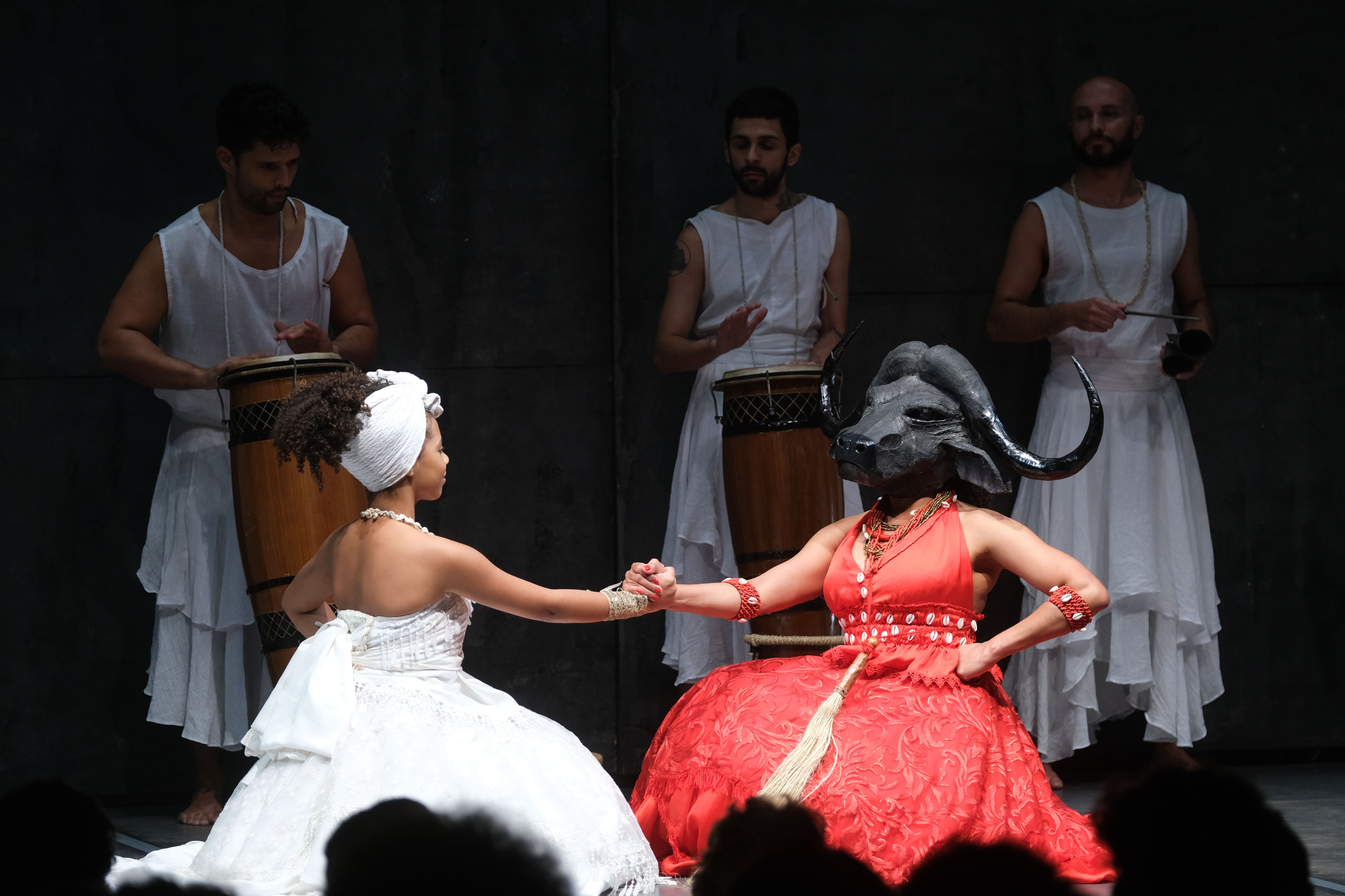 A imagem mostra uma cena teatral com cinco performers em um palco. No centro, em primeiro plano, duas mulheres est&atilde;o ajoelhadas e de m&atilde;os dadas, olhando uma para a outra. A mulher da esquerda tem pele escura, cabelo crespo preso em um turbante branco, e veste um vestido branco volumoso e um colar de p&eacute;rolas. A mulher da direita, tamb&eacute;m de pele escura, veste um vestido vermelho volumoso, um colar e pulseiras vermelhas, e usa uma m&aacute;scara de cabe&ccedil;a de b&uacute;falo realista, com chifres pretos e orelhas.

Ao fundo, h&aacute; tr&ecirc;s homens de pele clara, todos vestindo t&uacute;nicas brancas e aparentemente sem camisa por baixo. Os dois homens nas laterais est&atilde;o tocando atabaques, tambores de madeira, enquanto o homem do centro observa a cena. O cen&aacute;rio do palco &eacute; escuro e minimalista, com uma parede cinza ou preta ao fundo. A ilumina&ccedil;&atilde;o &eacute; forte e focada nos int&eacute;rpretes, destacando as cores das roupas e a intensidade da cena.
