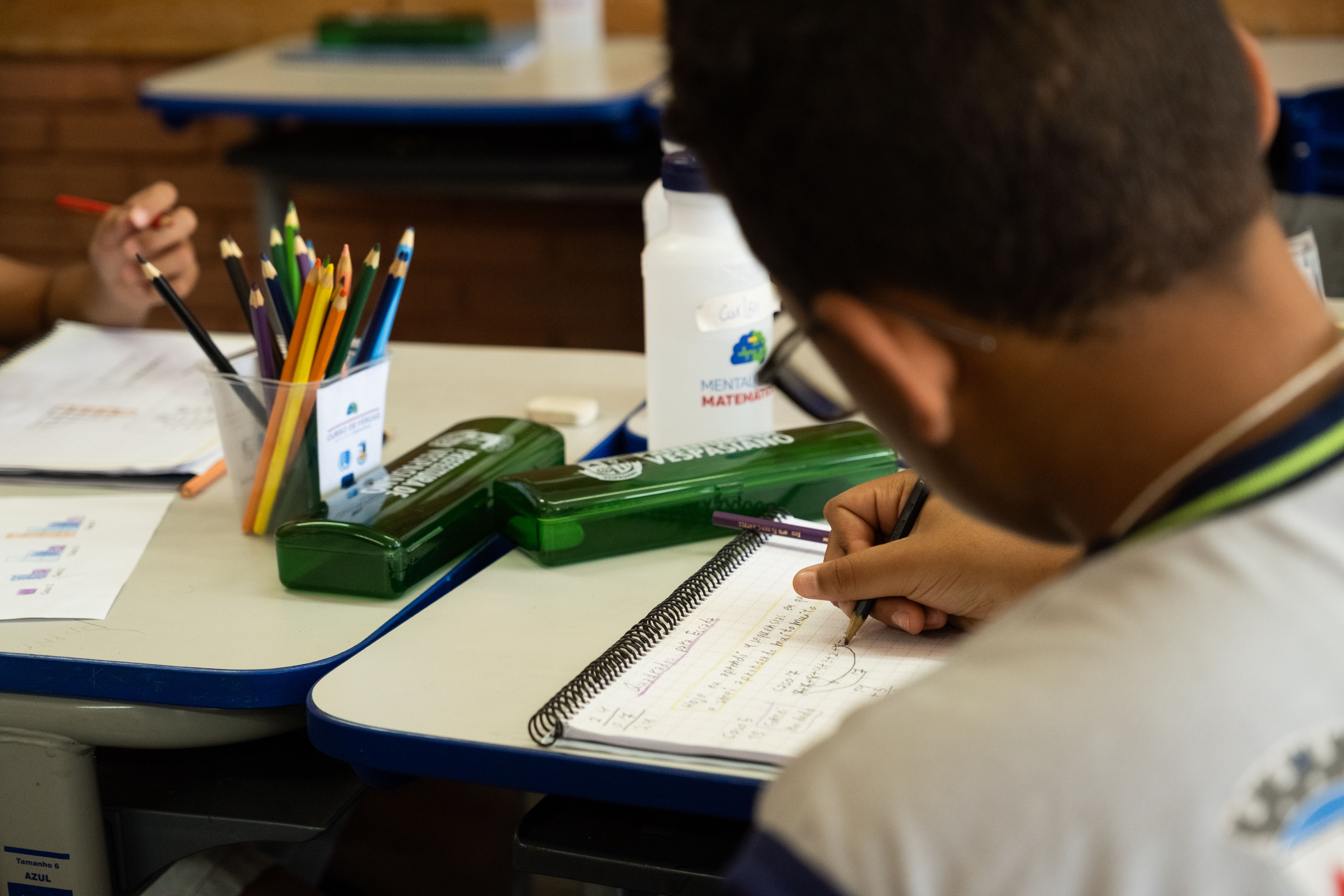 A foto &eacute; um close-up de um aluno em uma sala de aula, visto por cima do ombro. O aluno, com cabelo escuro e camisa clara com detalhes em azul e verde, est&aacute; sentado em uma carteira escolar e escrevendo em um caderno espiral com a m&atilde;o direita. No caderno, h&aacute; anota&ccedil;&otilde;es e desenhos.
Na carteira &agrave; frente do aluno, ou na mesma carteira, h&aacute; um copo com v&aacute;rios l&aacute;pis de cor de diferentes cores, um estojo verde escuro e uma garrafa de &aacute;gua branca com um r&oacute;tulo azul. Ao fundo, as m&atilde;os de outro aluno s&atilde;o vis&iacute;veis, tamb&eacute;m escrevendo. O ambiente &eacute; de sala de aula, com mesas e cadeiras claras. A ilumina&ccedil;&atilde;o &eacute; natural e foca na atividade de estudo.