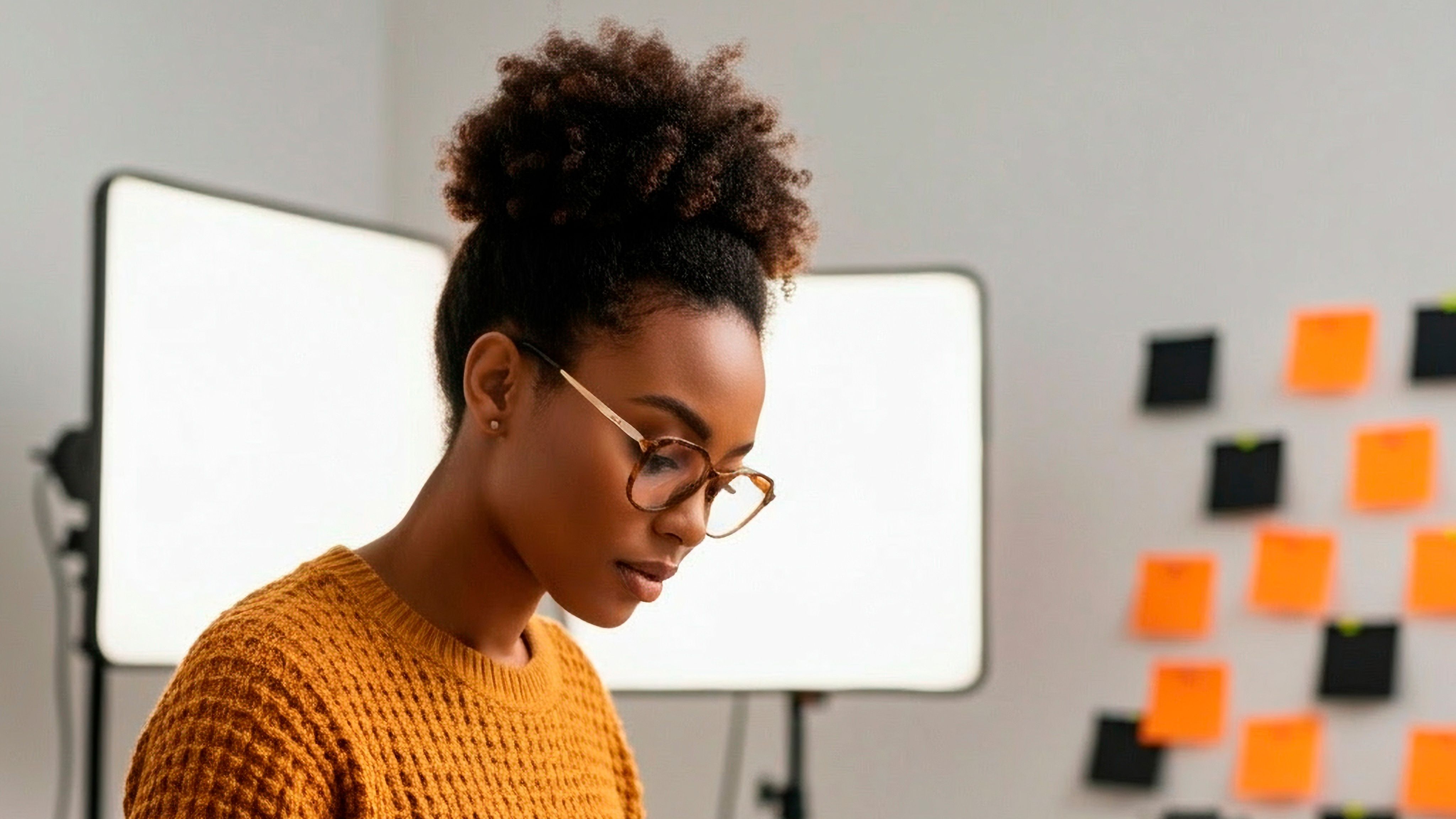 Mulher negra de óculos e cabelo crespo preso em coque alto aparece concentrada, usando suéter mostarda. Ao fundo, há painéis de luz e uma parede com post-its coloridos. A cena sugere ambiente de trabalho criativo ou estudo.