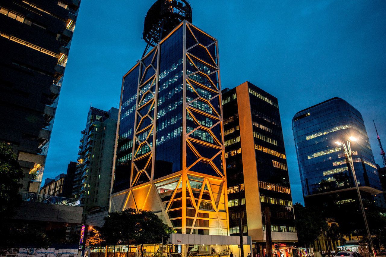 Fotografia tirada à noite, mostrando o prédio do Itaú Cultural do outro lado da avenida. Há uma luz amarela no prédio. Outros edifícios aparecem ao redor.