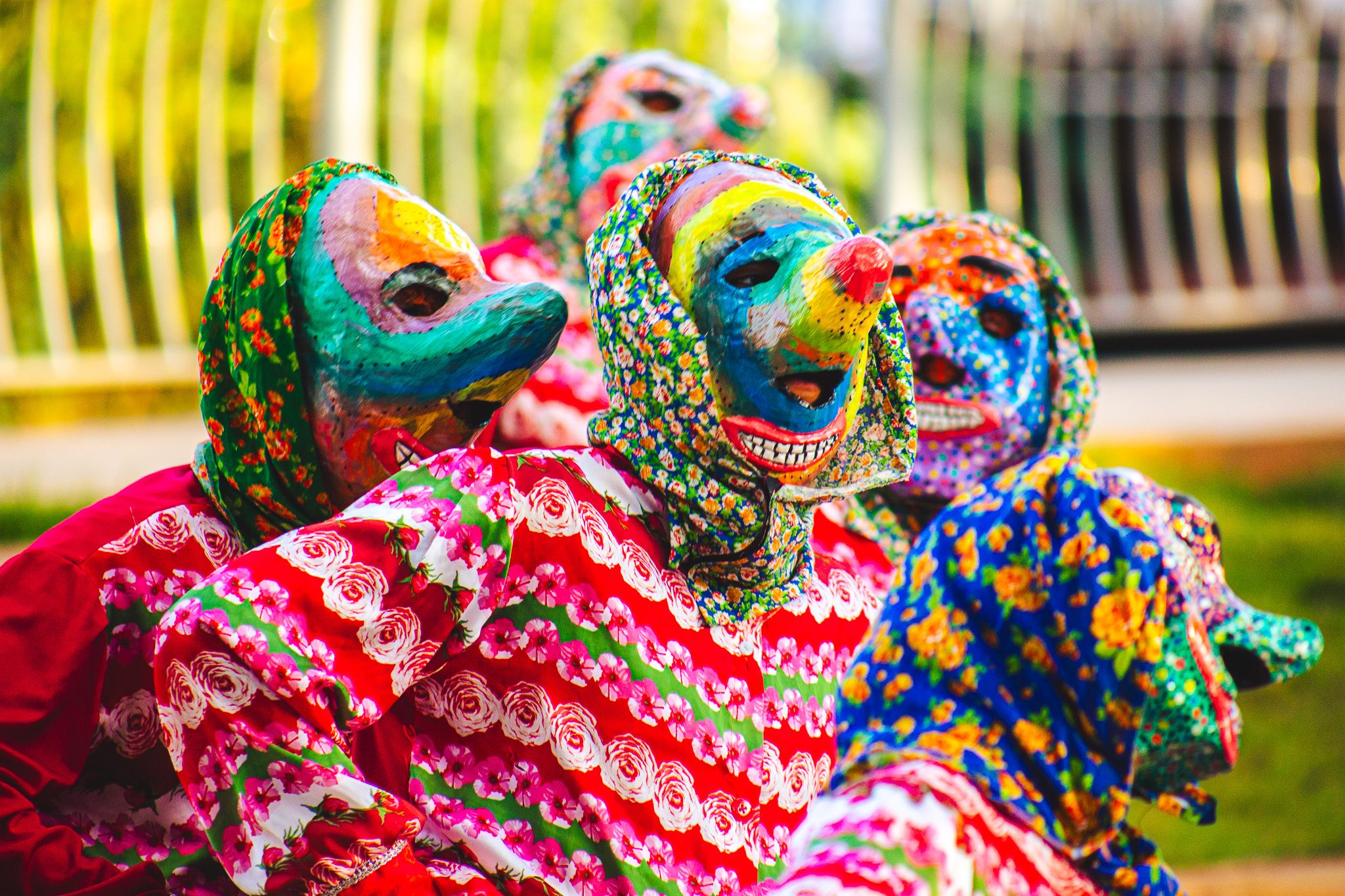 Cinco figuras com máscaras coloridas, que se assemelham a bichos, formam uma fila. Eles vestem capuzes e roupas floridas