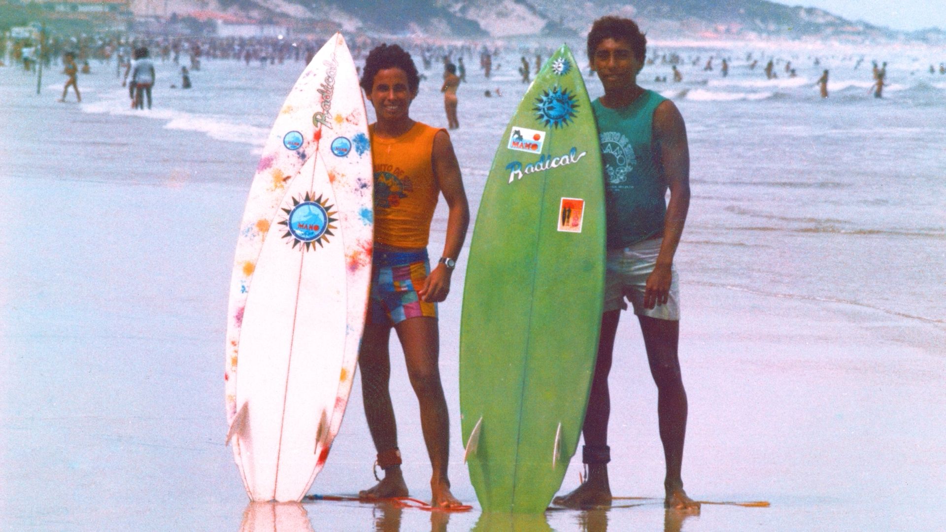 Foto com aspecto antigo de dois jovens surfistas sorrindo em uma praia lotada. Em pé na areia molhada, eles seguram suas pranchas na vertical, que são coloridas e têm adesivos.