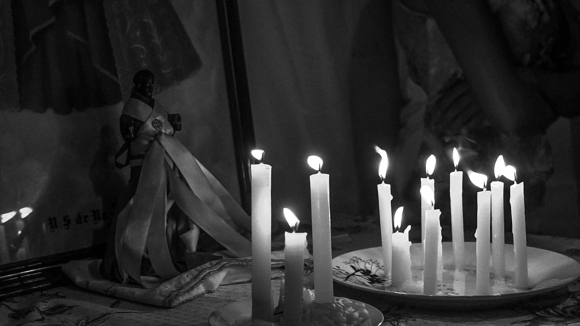 Foto em preto e branco de um altar à luz de velas. As chamas de várias velas brancas, dispostas em pratos, iluminam um ambiente escuro. Ao fundo, vê-se a estátua de um santo negro.