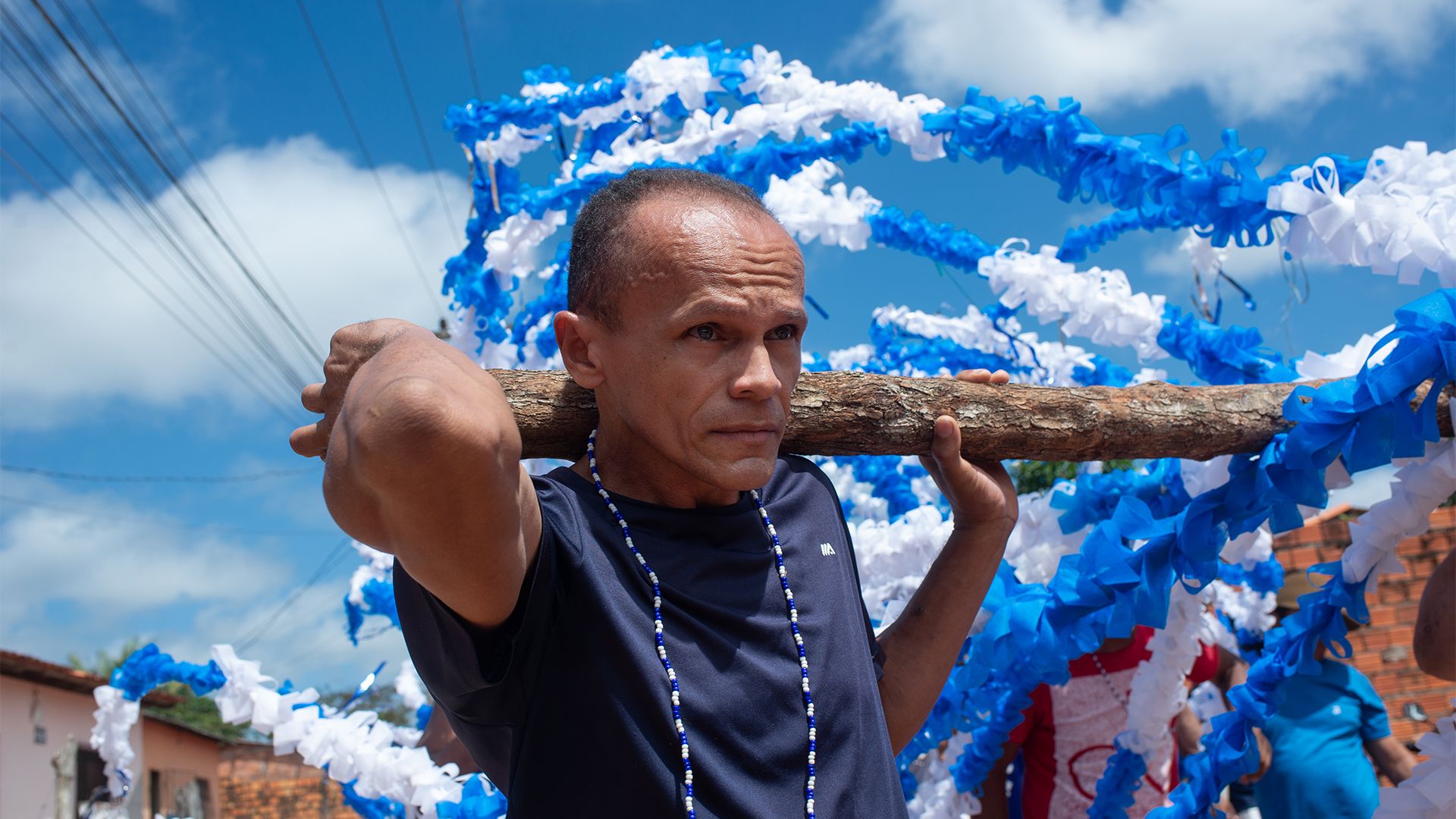 Em um cortejo de rua, sob um céu azul, um homem com expressão concentrada carrega um mastro de madeira nos ombros. O mastro é enfeitado com longas fitas de papel azuis e brancas.