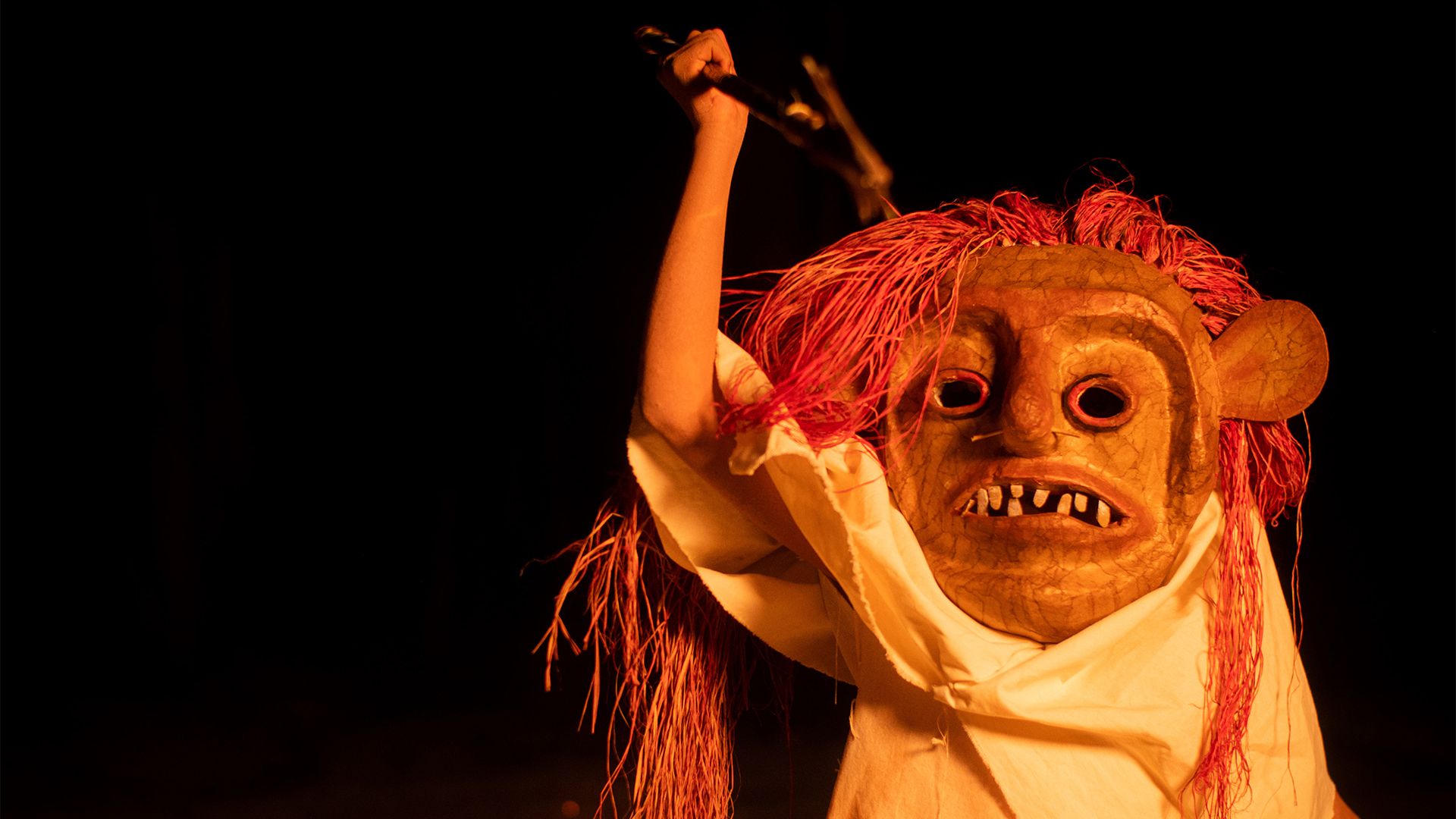Pessoa mascarada em um ritual noturno, iluminada por uma fogueira. A máscara, com cabelos vermelhos e dentes à mostra, tem um ar ameaçador. A figura, com túnica clara, ergue uma lança sobre a cabeça.