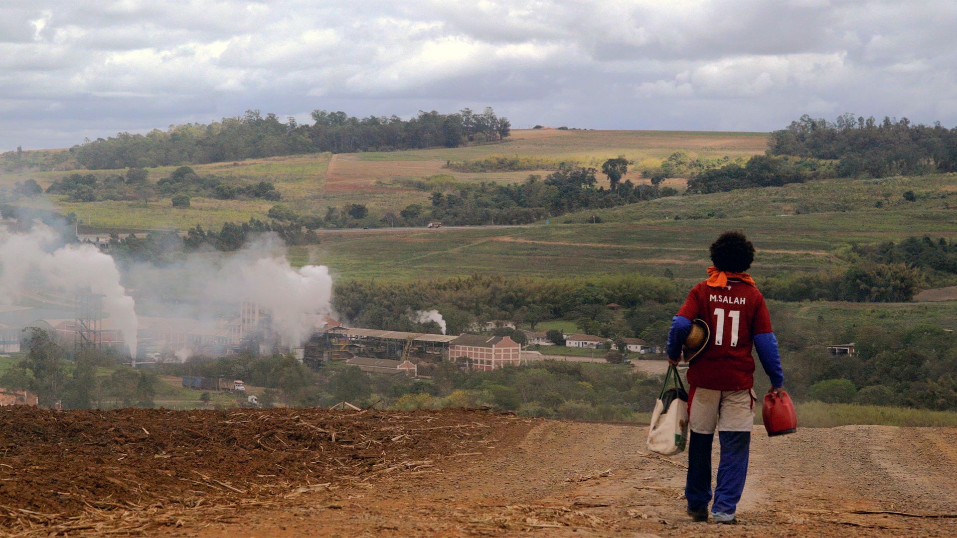 Homem de costas com camisa de futebol de M. Salah desce uma estrada de terra. Ao fundo, paisagem rural com morros e uma usina com chaminés soltando fumaça branca sob céu nublado.