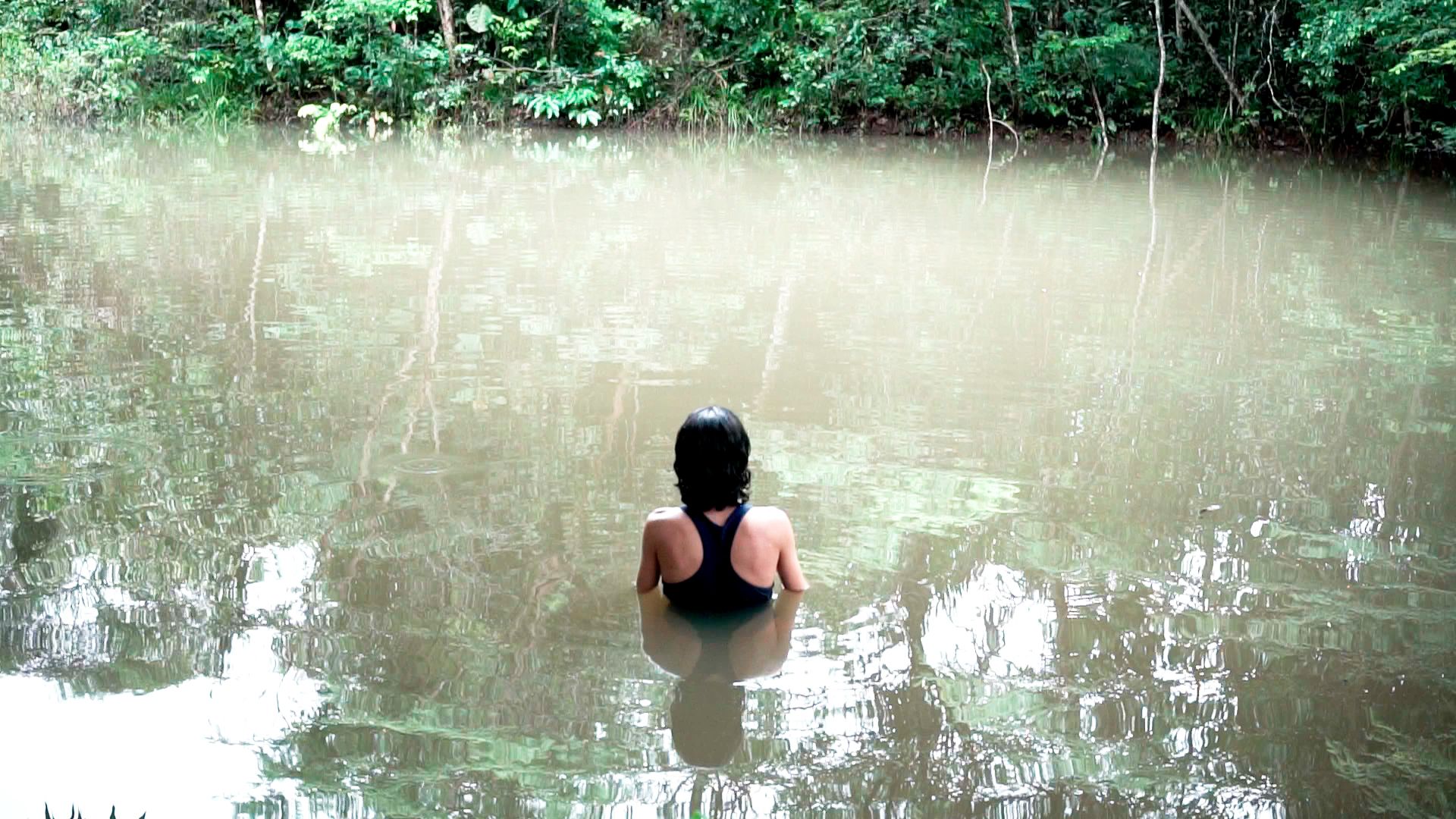 De costas, uma pessoa de cabelos escuros está imersa até os ombros em um rio de águas calmas. A margem ao fundo é coberta por uma floresta densa, cujo verde se reflete na superfície da água.