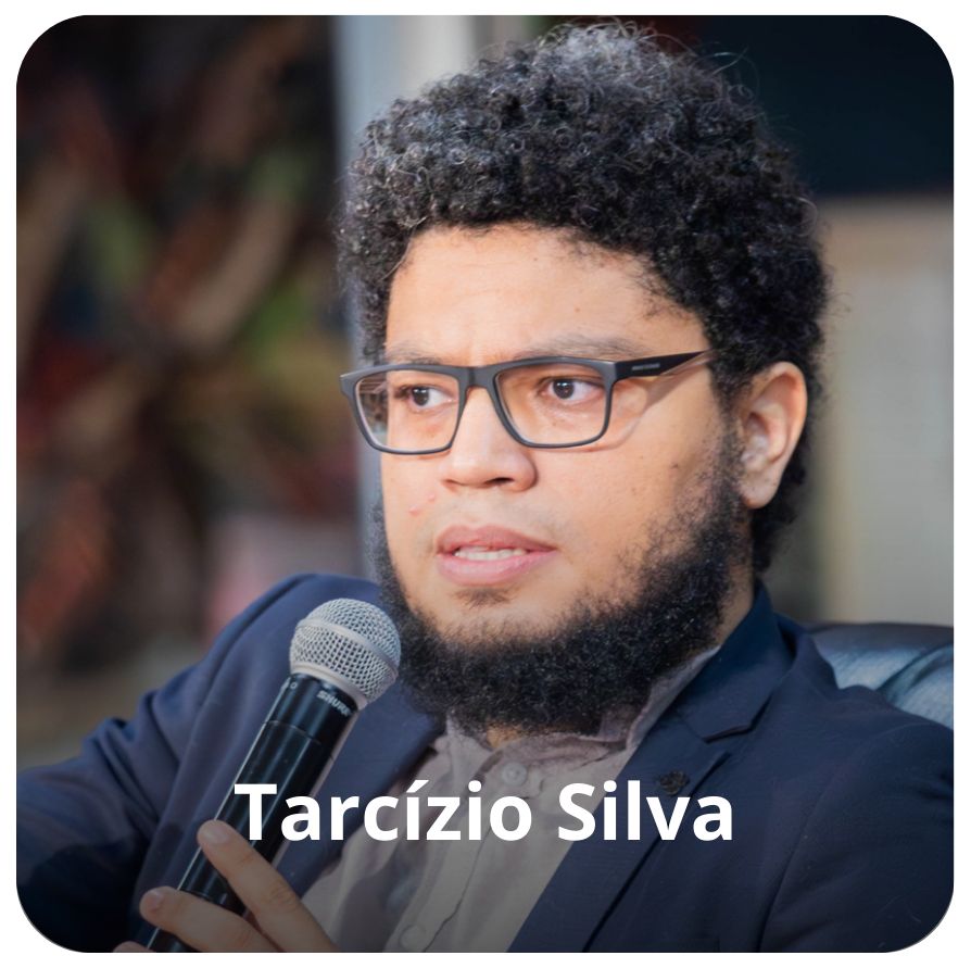 Um homem negro, de pele clara, de barba, cabelos crespos, curtos e preto. Ele usa &oacute;culos, uma paleto na cor azul escuro e segura um microfone enquanto fala e olha em dire&ccedil;&atilde;o ao p&uacute;blico 