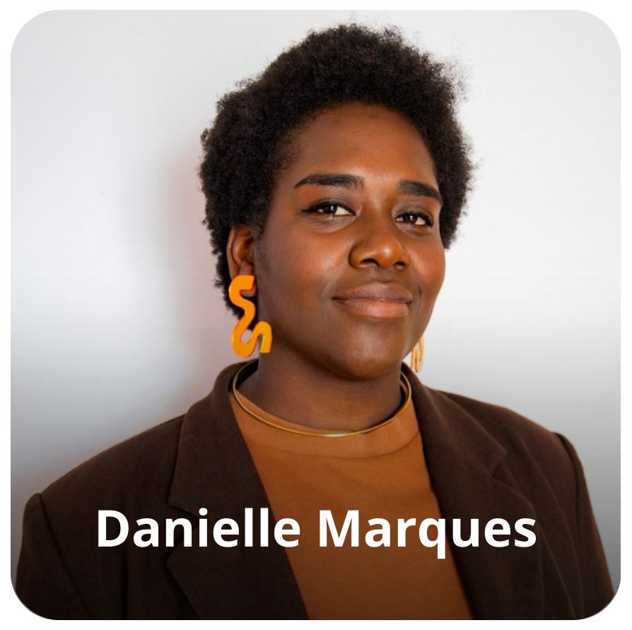 Uma mulher negra, de cabelos crespos, curtos e preto. Ela usa um brinco amarelo, veste roupas em tons terrosos e est&aacute; olhando em dire&ccedil;&atilde;o a c&acirc;mera 