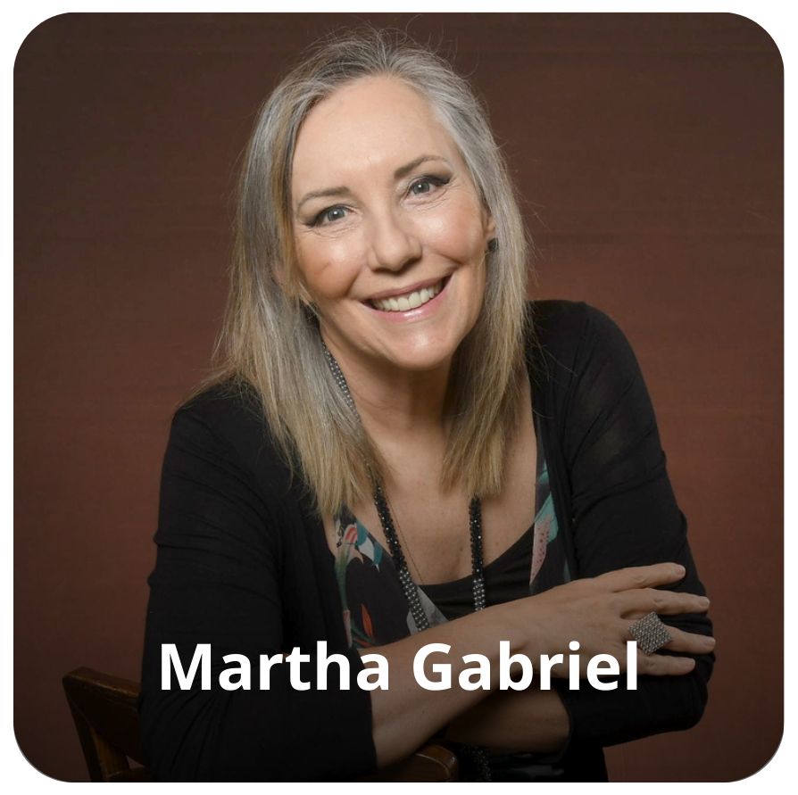 Uma mulher branca, de cabelos brancos nas altura dos ombros. Ela est&aacute; sorrindo em dire&ccedil;&atilde;o a c&acirc;mera. Ela veste uma blusa de mangas longas na cor preta e est&aacute; sentada em uma caderia marrom 