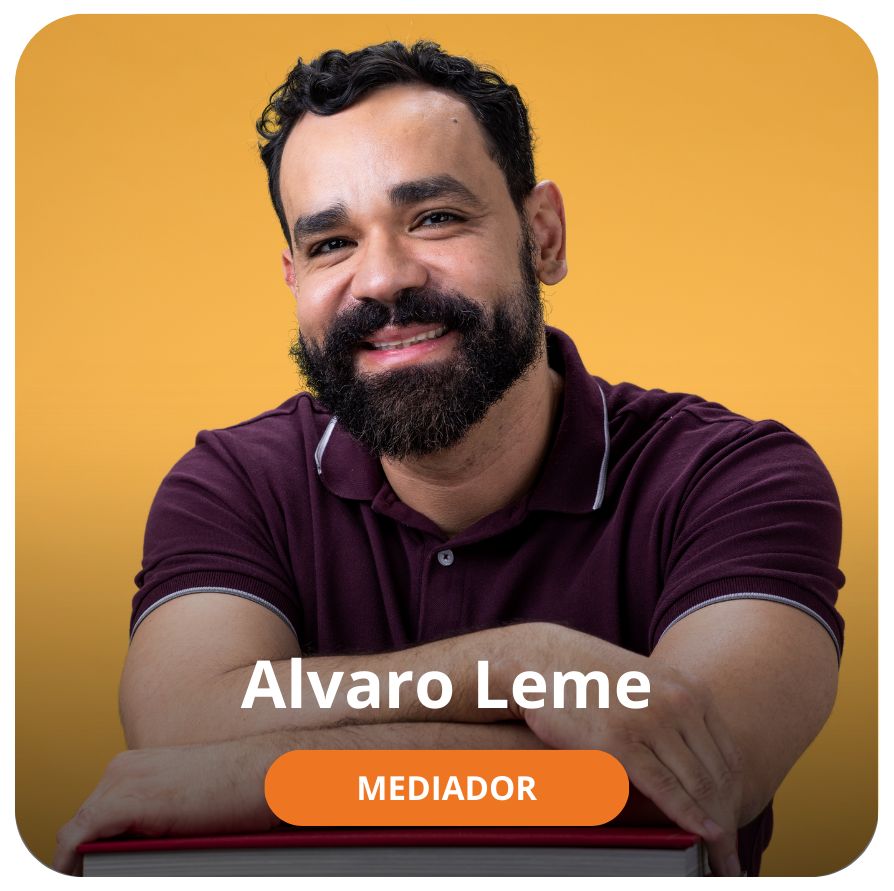 Um homem branco, jovem, de cabelos e barba curta e preta. Ele est&aacute; sorrindo, veste uma camisa na cor roxa e est&aacute; com os bra&ccedil;os apoiados em alguns livros. O fundo da foto &eacute; na cor laranja