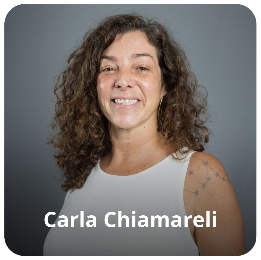 Uma mulher branca, de cabelos ondulados na altura dos ombros. Ela est&aacute; ventido uma camiseta branca e est&aacute; sorrindo. Atr&aacute;s dela est&aacute; um fundo e na cor cinza.