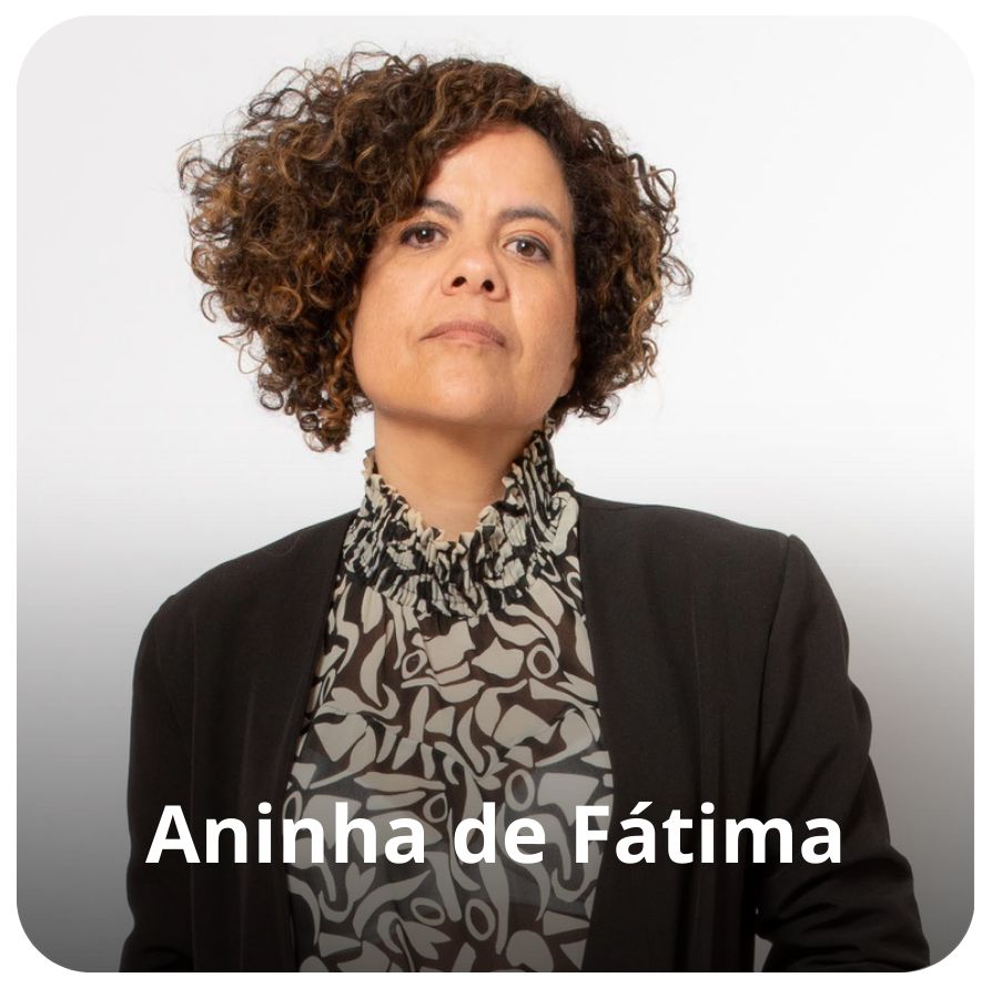 Uma mulher negra de pele clara, cabelos curtos e cacheados. Ela veste uma camisa com detalhes em preto e branco e um blazer preto. 