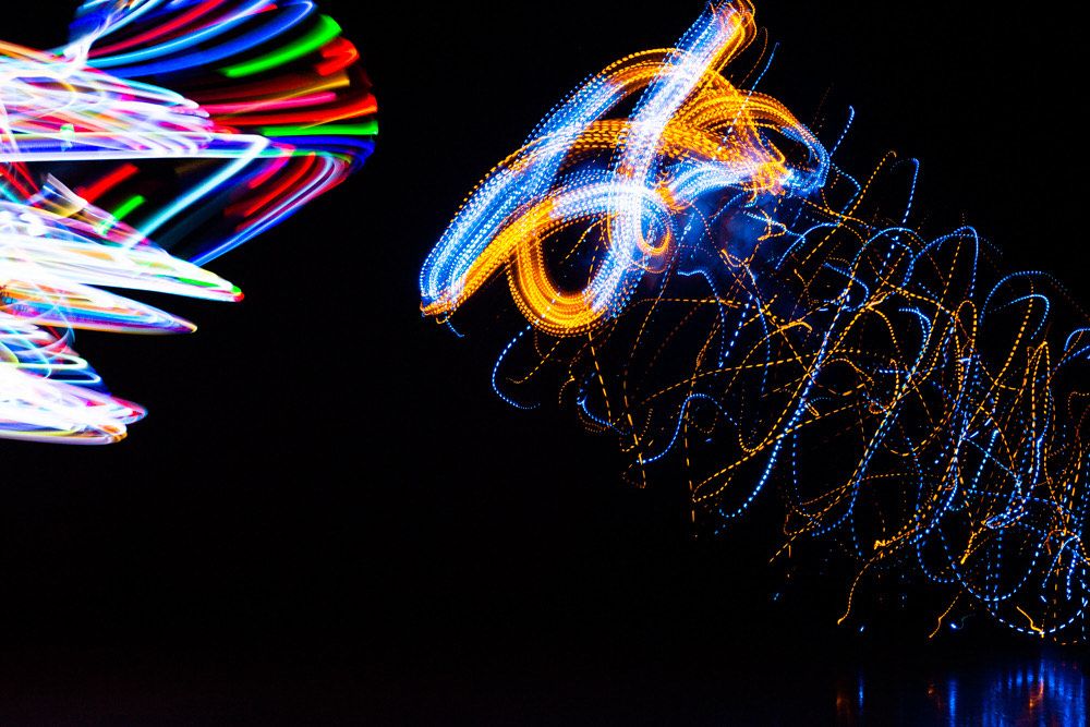 Fotografia abstrata em fundo preto. Rastros de luz vibrantes, em azul, laranja e outras cores, formam traços sinuosos e rabiscos no ar. As formas entrelaçadas criam uma intensa sensação de movimento, velocidade e energia.