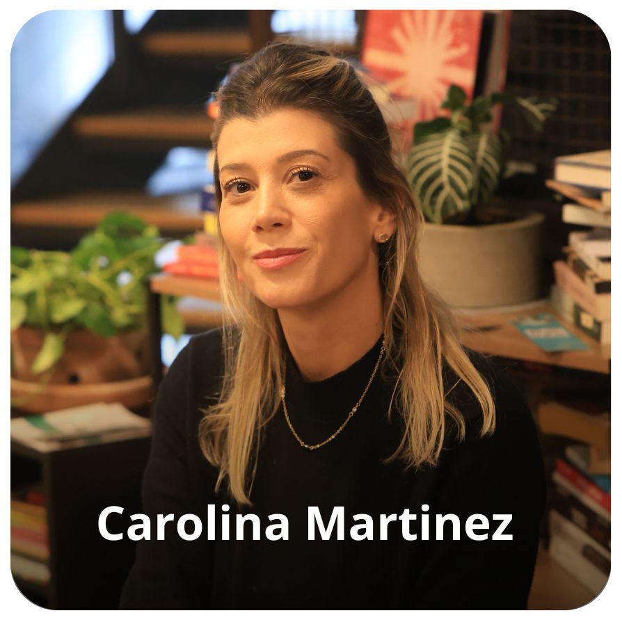 Uma mulher branca, de cabelo preto com mechas loiras na altura dos ombros. Ela veste uma blusa de mangas longas preta. Ao fundo est&atilde;o alguns livros e plantas. 