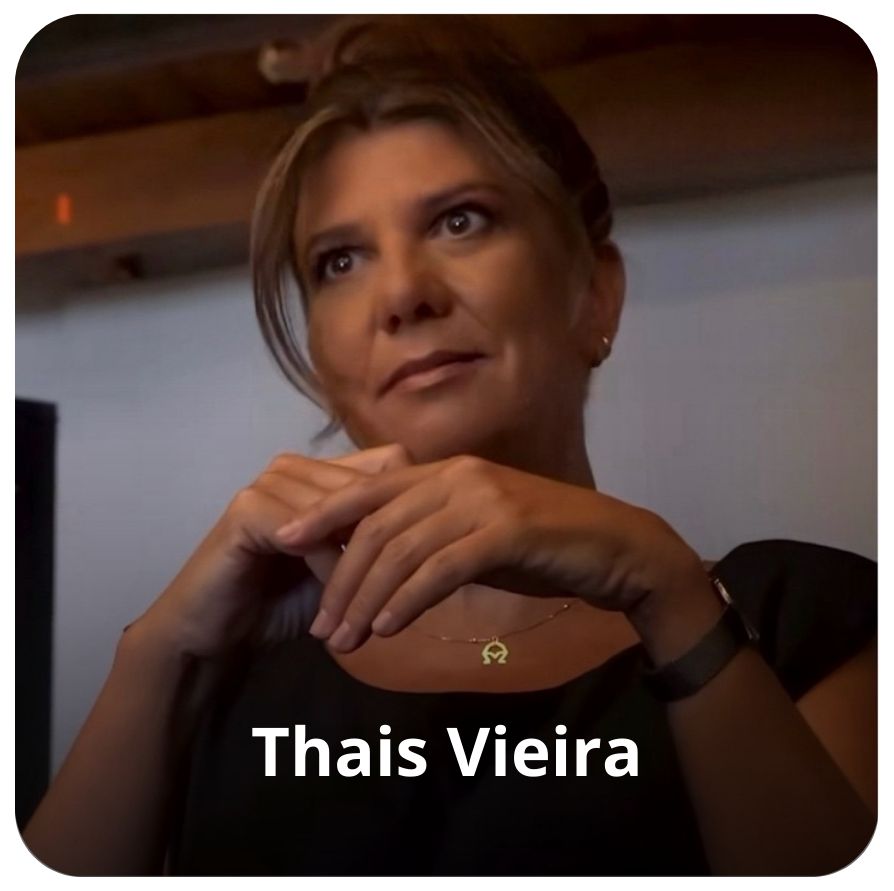 
A imagem mostra uma mulher  loira com mechas, sentada e olhando para o lado esquerdo da foto com uma express&atilde;o pensativa. Ela est&aacute; com as m&atilde;os entrela&ccedil;adas sobre o colo ou uma mesa, e usa uma blusa preta.