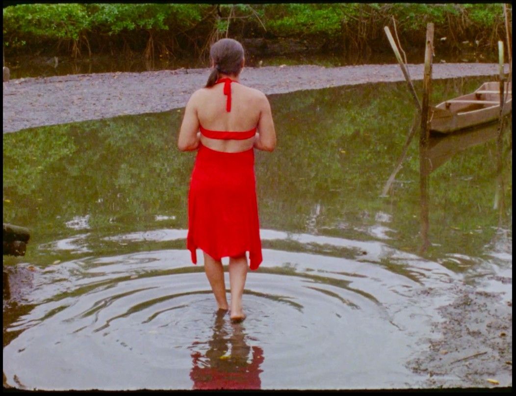 Carla da Victória está de costas, no rio. Ela usa um vestido vermelho e está descalça. Ao fundo, há árvores e uma canoa.