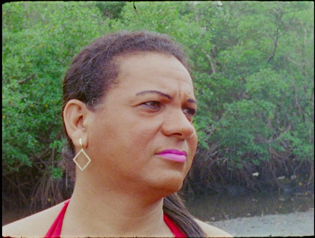 Carla da Victória está séria. Ela usa batom rosa e brincos dourados. Atrás dela, há árvores.