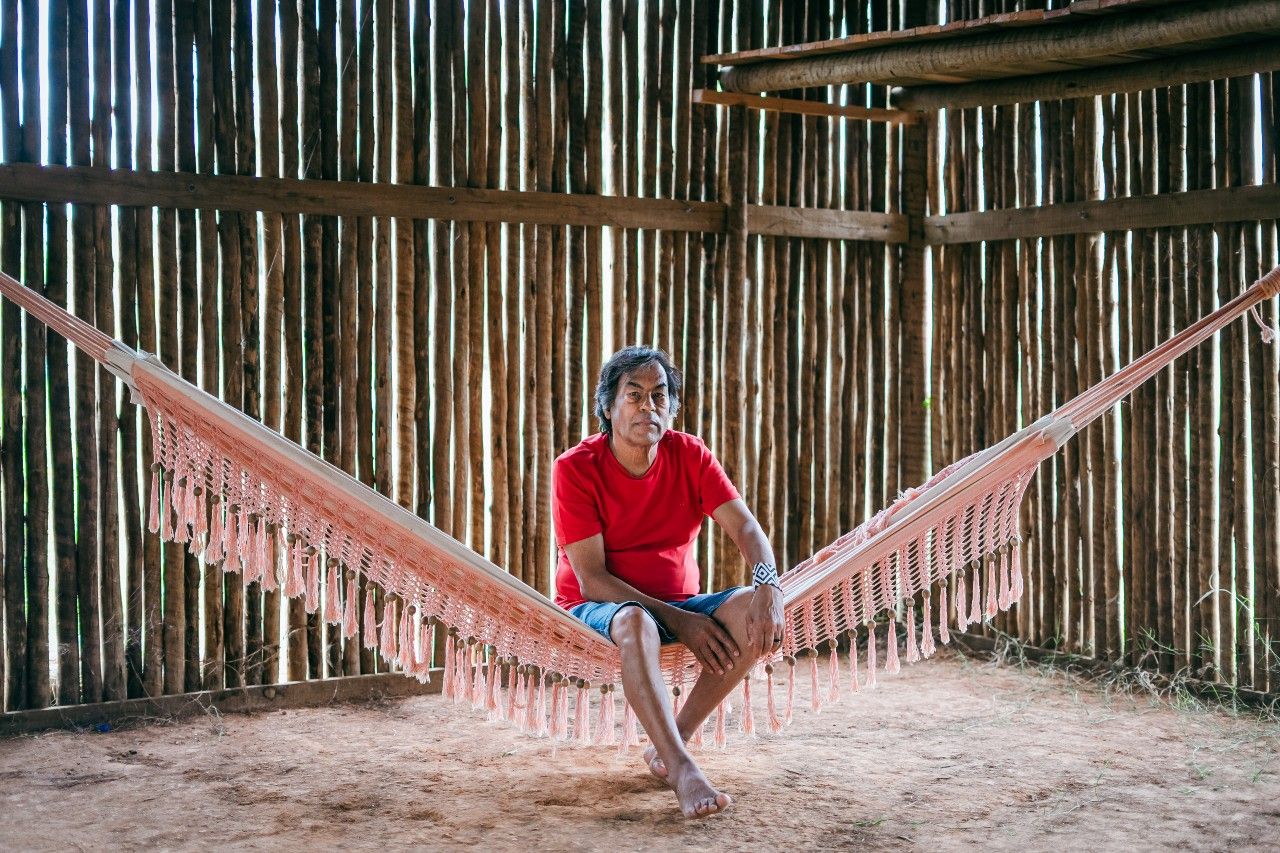 Ailton está sentado em uma rede e olha para a câmera. É um homem indígena de pele escura e cabelos negros.