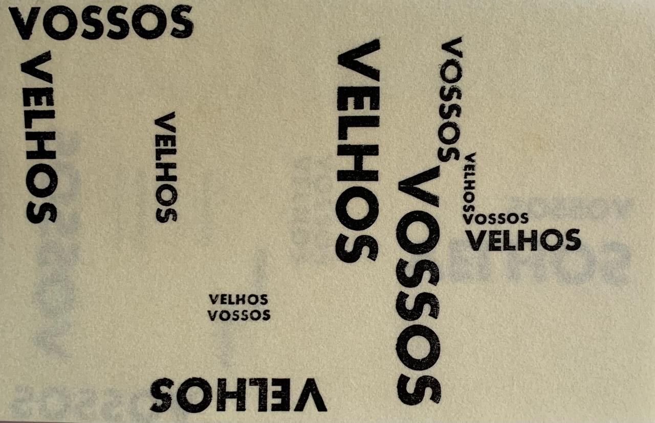 A imagem apresenta um fundo bege com palavras pretas dispostas de forma artística. As palavras "vossos" e "velhos" aparecem repetidamente em diferentes tamanhos e orientações. Algumas palavras estão na vertical, outras na horizontal, e algumas estão giradas em ângulos variados. O padrão visual parece brincar com a repetição e a disposição espacial das palavras. O contraste entre o fundo claro e as letras escuras torna o texto bem legível.