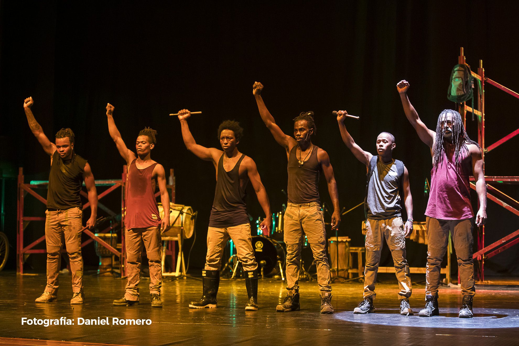 Seis homens negros estão alinhados em um palco, com os braços esquerdos erguidos e os punhos cerrados. Eles vestem camisetas regatas e calças, e alguns seguram baquetas. Ao fundo, veem-se instrumentos de percussão e uma estrutura de andaime. A expressão deles é de força e seriedade.