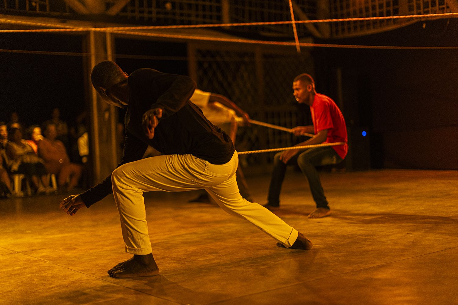 Em um ambiente com luz quente e amarelada, três homens negros estão em um palco. Um deles, em primeiro plano, veste calça clara e blusa escura e está agachado em uma pose de ação. Ao fundo, outro homem de camisa vermelha está sentado no chão, puxando com força uma corda branca e grossa. O terceiro homem, mais distante que o de vermelho, está parcialmente bloqueado pelo primeiro homem.