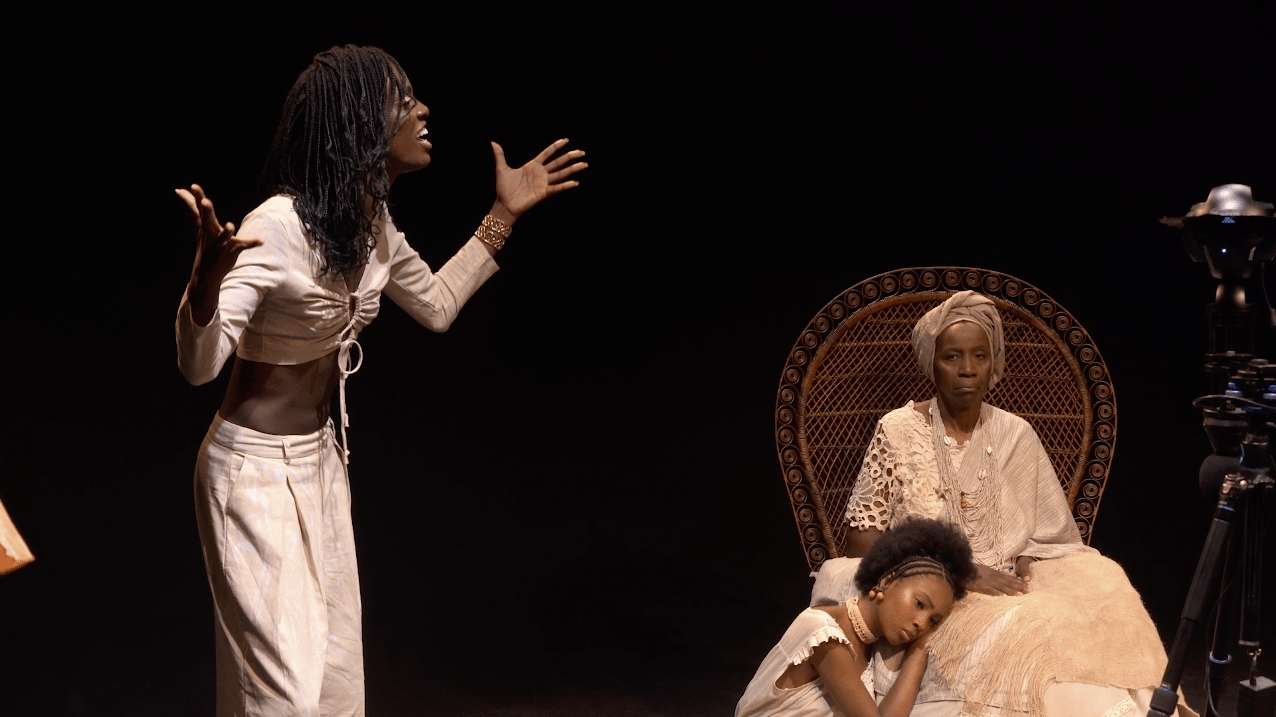 No palco, três mulheres negras, todas vestidas com roupas brancas. Uma está em pé. Outra, a mais velha, encontra-se sentada em uma cadeira. E a terceira aparece sentada no chão, com a cabeça apoiada no colo da mais velha. Na imagem, há ainda uma câmera em um tripé.