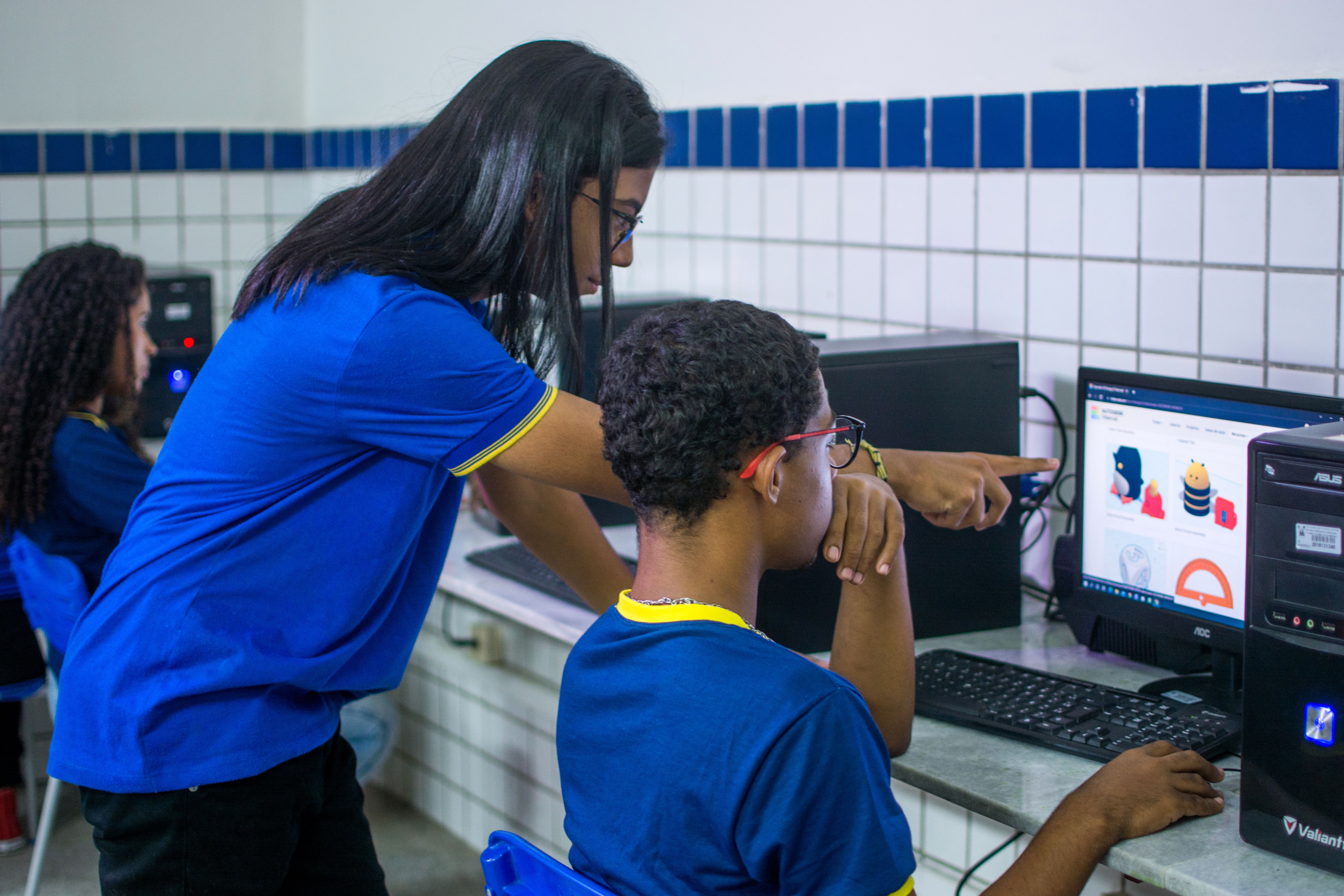 A foto mostra dois estudantes em um laborat&oacute;rio de inform&aacute;tica colaborando em uma tarefa. A estudante em p&eacute;, de uniforme azul, aponta para o monitor de um computador, enquanto o colega sentado observa a tela. O monitor exibe um software educacional com gr&aacute;ficos coloridos. O ambiente &eacute; caracterizado por azulejos brancos e uma faixa azul.