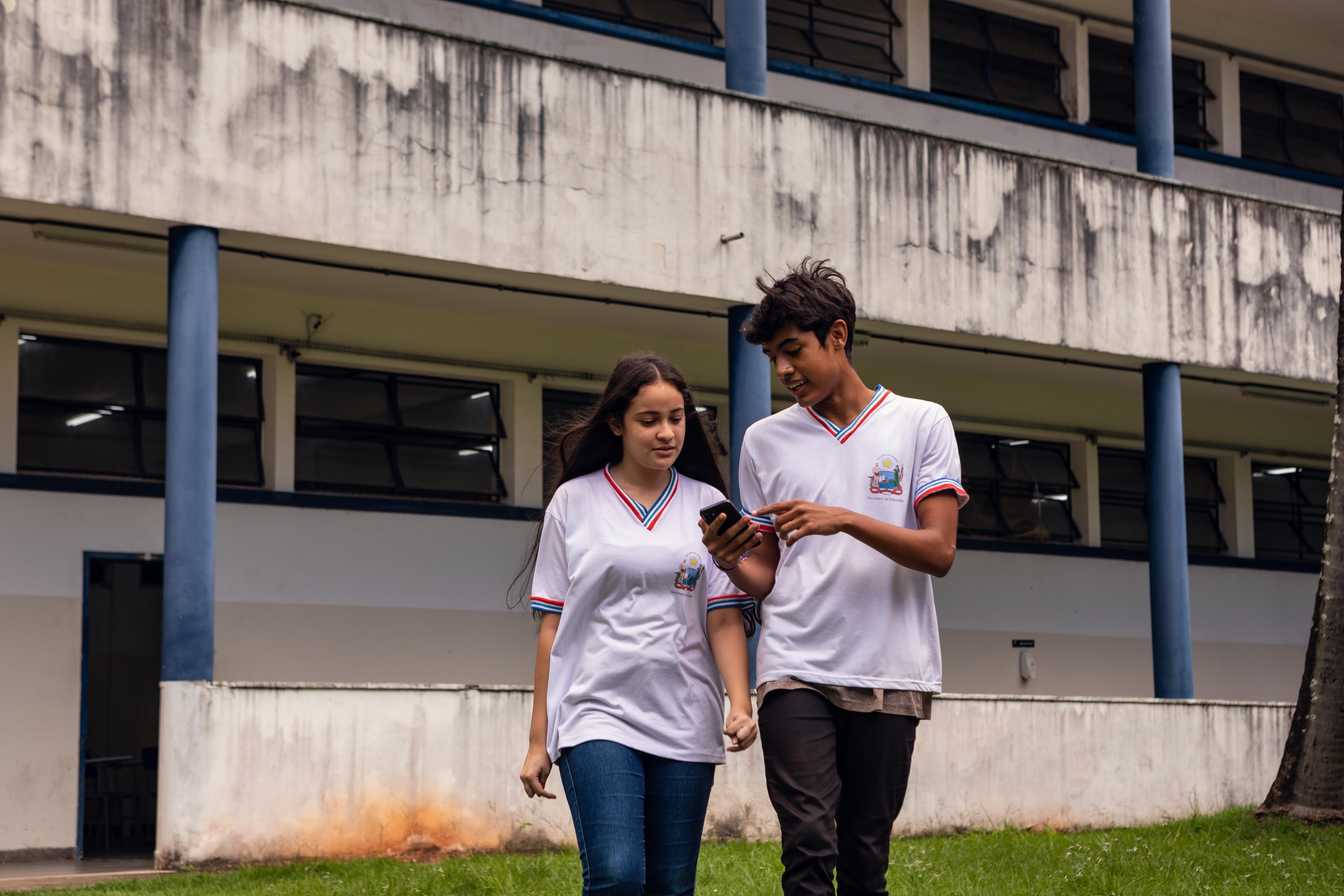 A foto retrata dois adolescentes, provavelmente estudantes em uniforme (camisetas brancas com detalhes em vermelho e azul), caminhando juntos em um gramado em frente a um pr&eacute;dio escolar de concreto com pilares azuis. O rapaz est&aacute; segurando um celular na m&atilde;o, e ambos est&atilde;o olhando para a tela enquanto caminham, sugerindo que est&atilde;o compartilhando conte&uacute;do ou se comunicando.