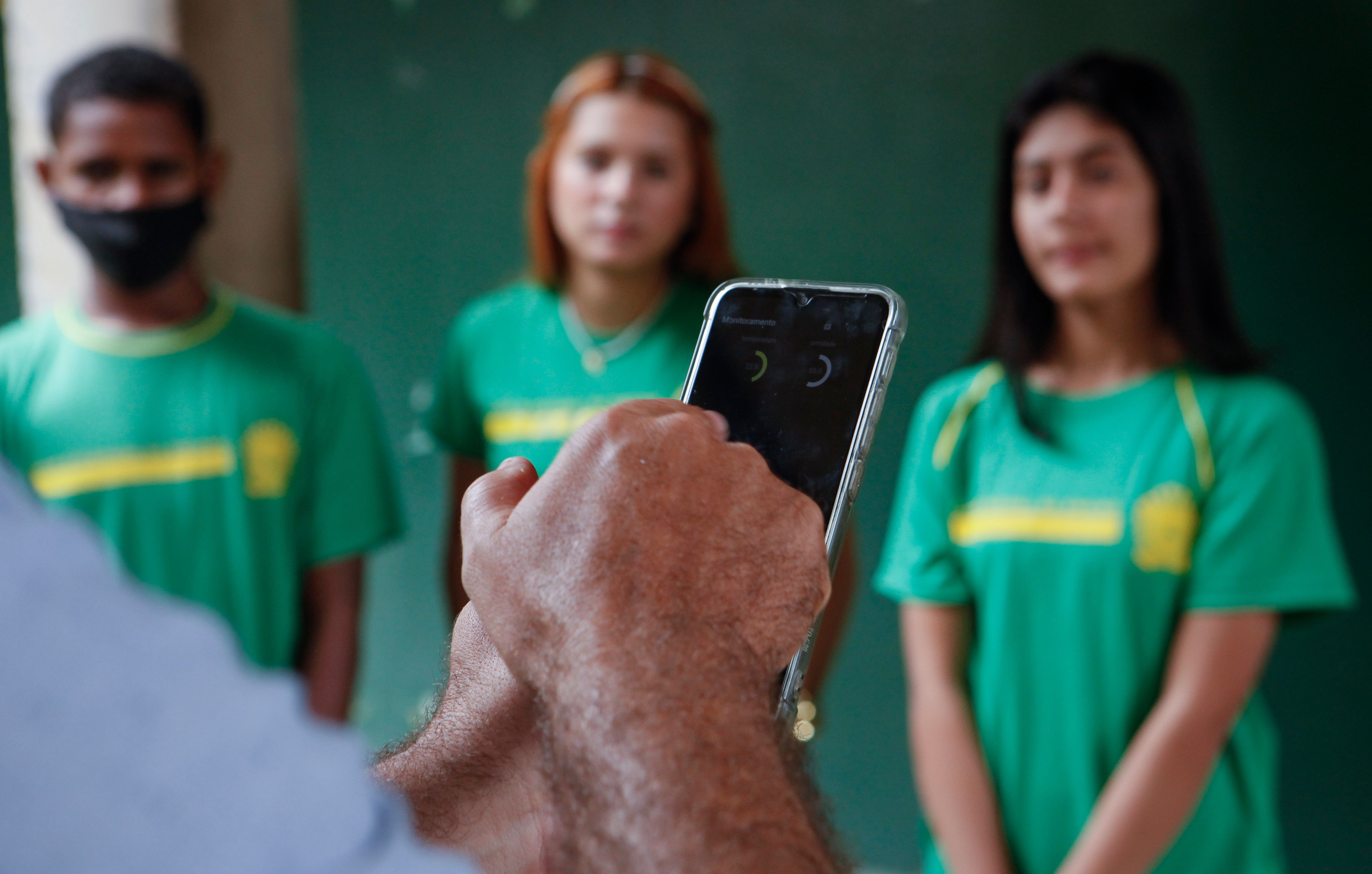 A foto &eacute; um close-up nas m&atilde;os de uma pessoa segurando um smartphone com a tela ligada, exibindo uma interface de monitoramento. Ao fundo, fora de foco, est&atilde;o tr&ecirc;s jovens em p&eacute;, todos vestindo uniformes escolares verdes com detalhes amarelos. A composi&ccedil;&atilde;o sugere que a pessoa em primeiro plano est&aacute; usando o aplicativo do celular para uma atividade ou avalia&ccedil;&atilde;o relacionada aos estudantes.