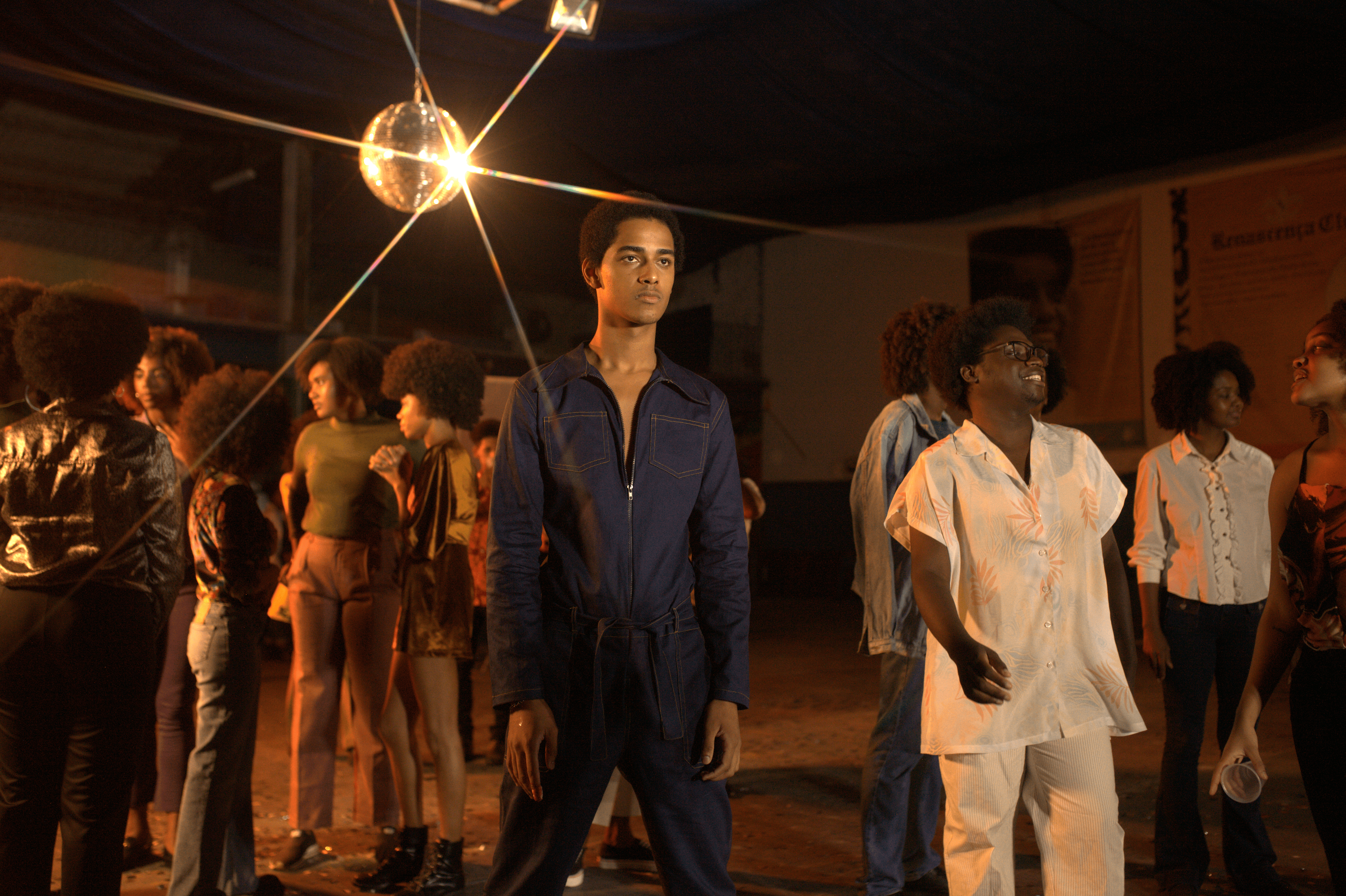 Ao centro, há um jovem negro que veste um macacão azul jeans e está sério, parado, fitando algo que não se vê. A luz do globo reflete por toda a pista e sobre ele também. Ao seu lado esquerdo, há um grupo de jovens, todos de black power com roupas que nos remetem aos anos 80. Ao seu lado direito, há também um grupo menor, mais ao fundo, no mesmo estilo, mas bem ao seu lado há outro jovem negro, vestido todo de branco e dançado mais próximo a ele. 