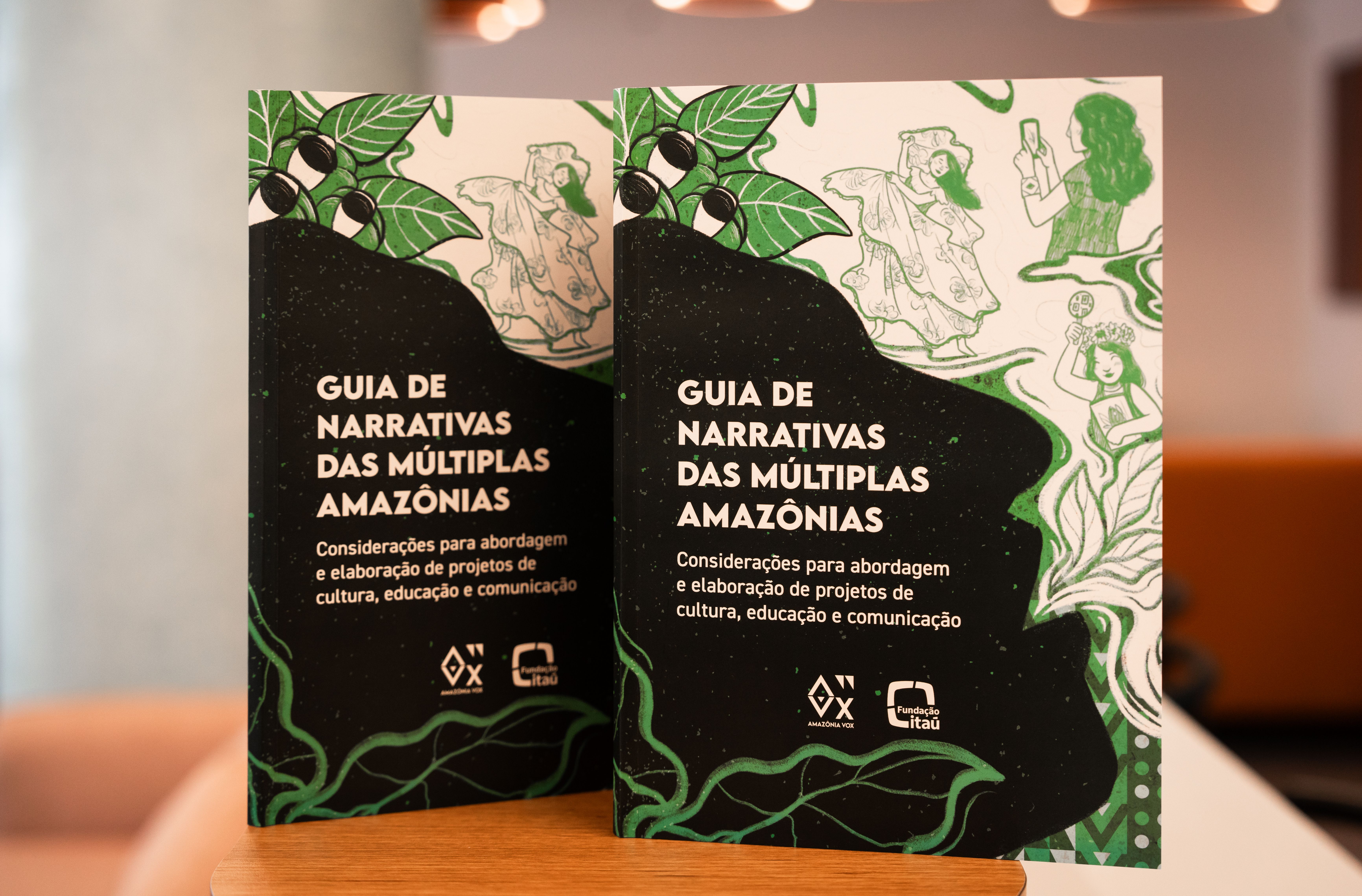 A imagem exibe dois exemplares id&ecirc;nticos de um livro em primeiro plano, posicionados lado a lado sobre uma superf&iacute;cie de madeira clara que pode ser uma mesa ou balc&atilde;o. A capa dos livros &eacute; predominantemente preta e verde, com o t&iacute;tulo em destaque: 