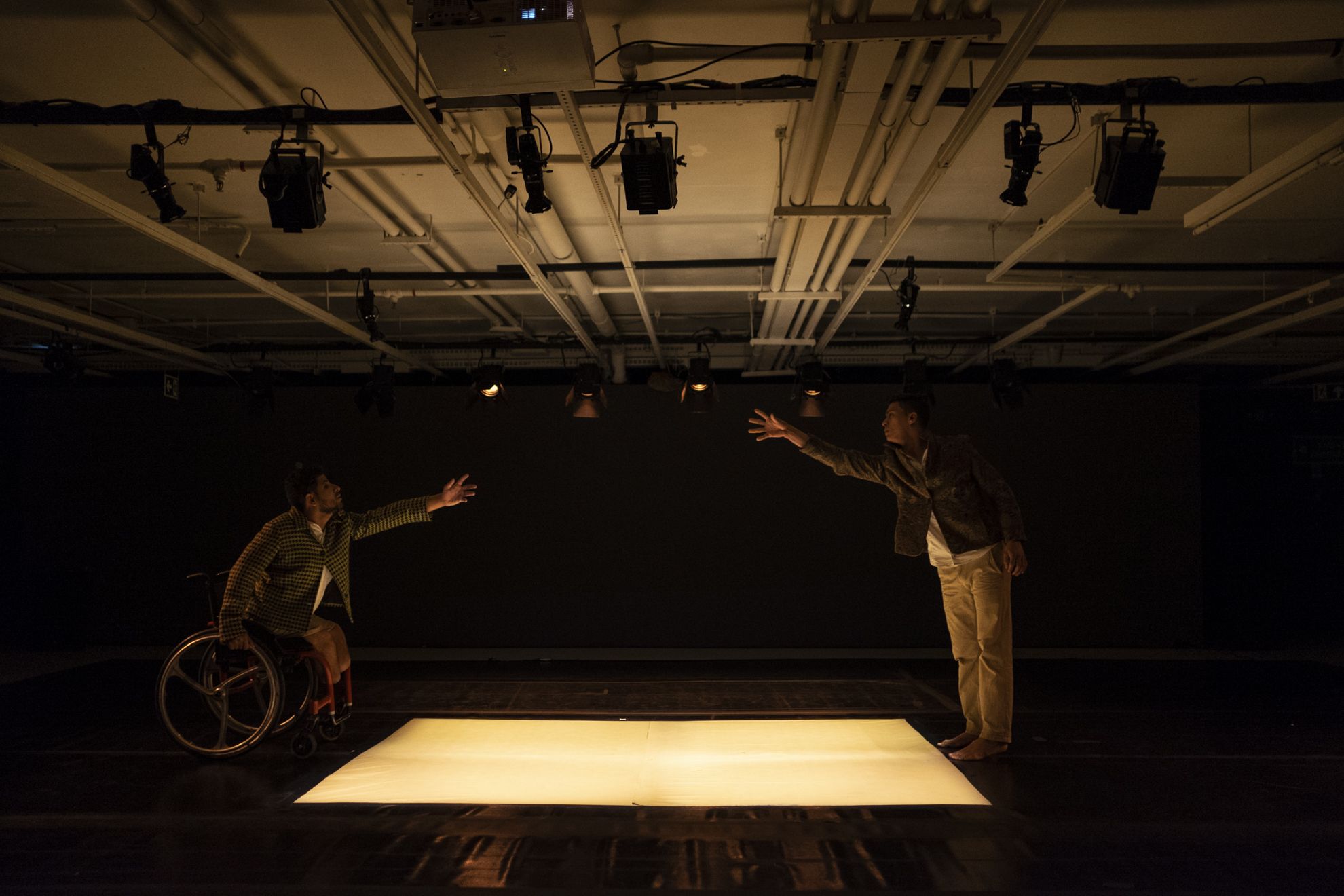 Dois homens se estendem um ao outro, separados por um retângulo de luz no chão escuro. A claridade do tapete branco parece respirar e pulsar luz, no centro da sala, cercada pela penumbra. À esquerda, o homem em cadeira de rodas projeta o corpo à frente, o braço esquerdo erguido em gesto de alcance. As pernas amputadas se revelam sob o short cor de areia. O homem na cadeira de rodas usa um casaco de lã quadriculado em tons de marrom e bege, abotoado sobre uma camisa branca. O semblante dele traz a urgência de quem quer tocar o outro. À direita, o segundo homem espelha o gesto: inclina-se para frente, a mão direita aberta no mesmo desejo de encontro. Usa roupas semelhantes, casaco, calça clara, e o rosto sem barba reflete o mesmo esforço silencioso. Ambos permanecem à margem da luz, sobre o piso negro que se confunde com o fundo da cena. Acima deles, o teto branco exibe um emaranhado geométrico de tubos e refletores que projetam uma iluminação amarelada e densa, como se o ar inteiro vibrasse com o impulso contido desse toque a distância. Dois homens se estendem um ao outro, separados por um retângulo de luz no chão escuro. A claridade do tapete branco parece respirar e pulsar luz, no centro da sala, cercada pela penumbra. À esquerda, o homem em cadeira de rodas projeta o corpo à frente, o braço esquerdo erguido em gesto de alcance. As pernas amputadas se revelam sob o short cor de areia. O homem na cadeira de rodas usa um casaco de lã quadriculado em tons de marrom e bege, abotoado sobre uma camisa branca. O semblante dele traz a urgência de quem quer tocar o outro. À direita, o segundo homem espelha o gesto: inclina-se para frente, a mão direita aberta no mesmo desejo de encontro. Usa roupas semelhantes, casaco, calça clara, e o rosto sem barba reflete o mesmo esforço silencioso. Ambos permanecem à margem da luz, sobre o piso negro que se confunde com o fundo da cena. Acima deles, o teto branco exibe um emaranhado geométrico de tubos e refletores que projetam uma iluminação amarelada e densa, como se o ar inteiro vibrasse com o impulso contido desse toque a distância.