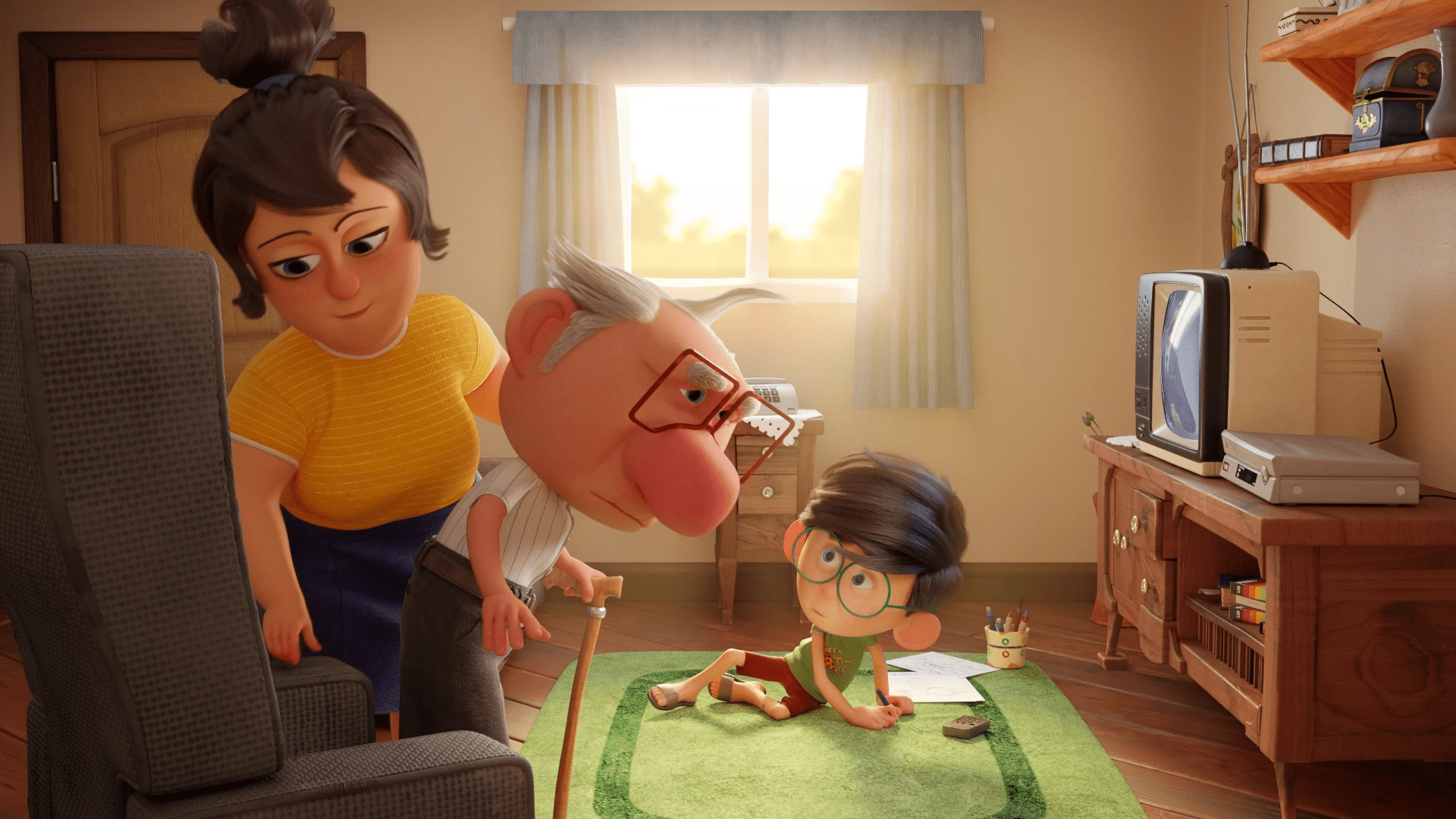 O avô está sentando na poltrona, com a ajuda da mãe. Do chão da sala, no tapete verde, o menino olha para os dois adultos. A avô usa bengala. No espaço, há também uma janela e uma TV de tubo.