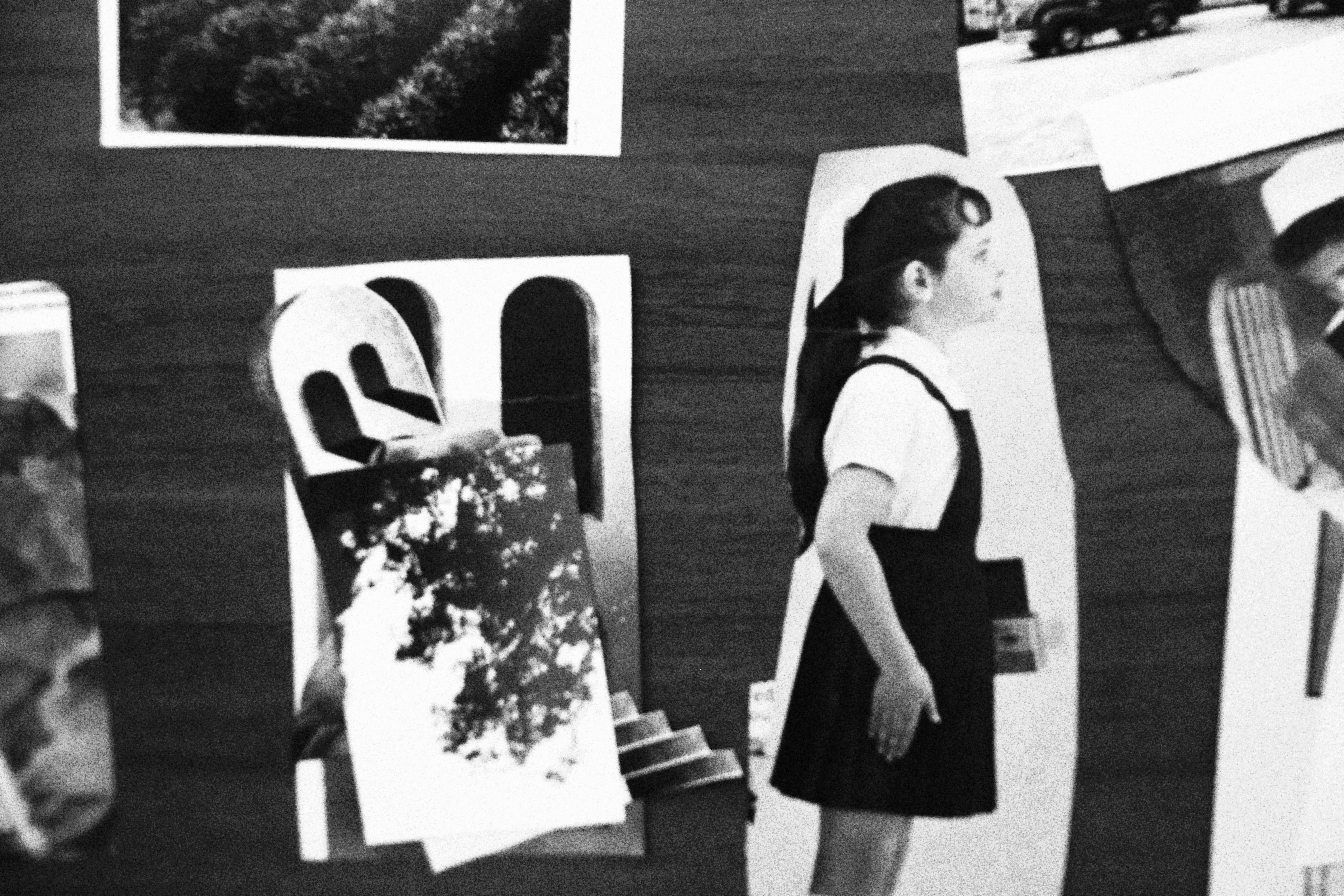 Uma foto granulada em preto e branco de uma colagem. O elemento central é o recorte de uma menina, vista de perfil, olhando para a direita. Ela usa um vestido escuro sobre uma camisa branca e tem o cabelo preso em um rabo de cavalo. À sua esquerda, há outro recorte com duas formas de arco, como janelas, sobreposto por uma foto texturizada de folhagem. No canto superior esquerdo, vê-se parte de uma foto de um campo. Uma foto granulada em preto e branco de uma colagem. O elemento central é o recorte de uma menina, vista de perfil, olhando para a direita. Ela usa um vestido escuro sobre uma camisa branca e tem o cabelo preso em um rabo de cavalo. À sua esquerda, há outro recorte com duas formas de arco, como janelas, sobreposto por uma foto texturizada de folhagem. No canto superior esquerdo, vê-se parte de uma foto de um campo.